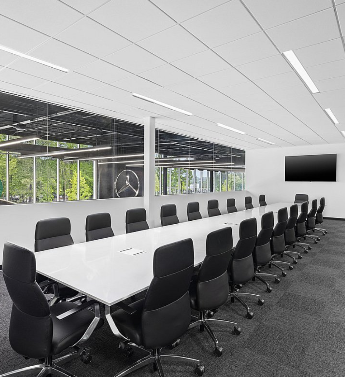 Modern conference room with a long white table surrounded by black office chairs, a large flat-screen TV on the white wall, and large windows showing green trees outside.