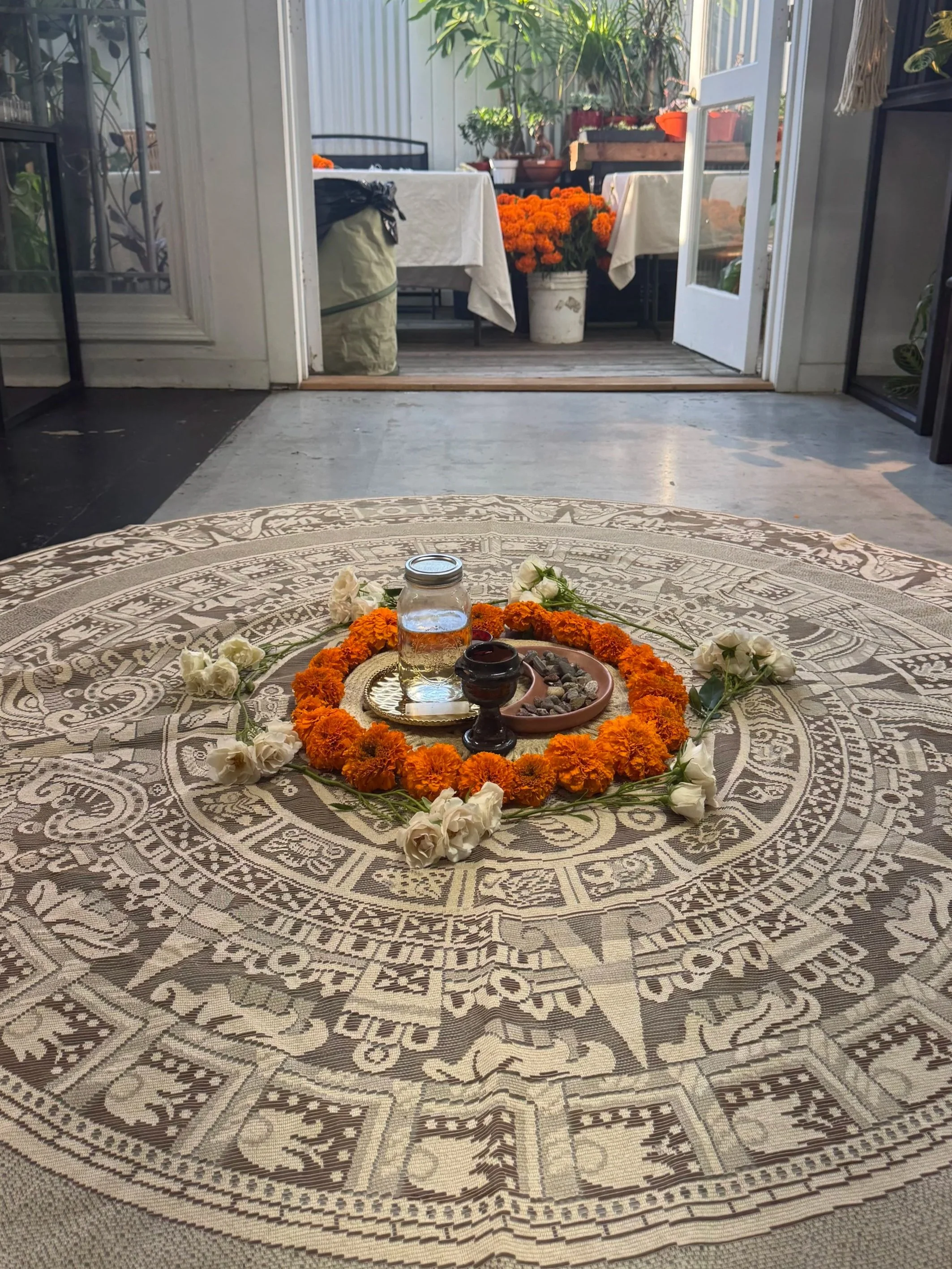 A round decorative tablecloth with intricate patterns and fringed edges, placed on a table with floral arrangements, candles, and a glass jar in the center. The background shows an open door leading to a patio area with potted plants and outdoor furniture.