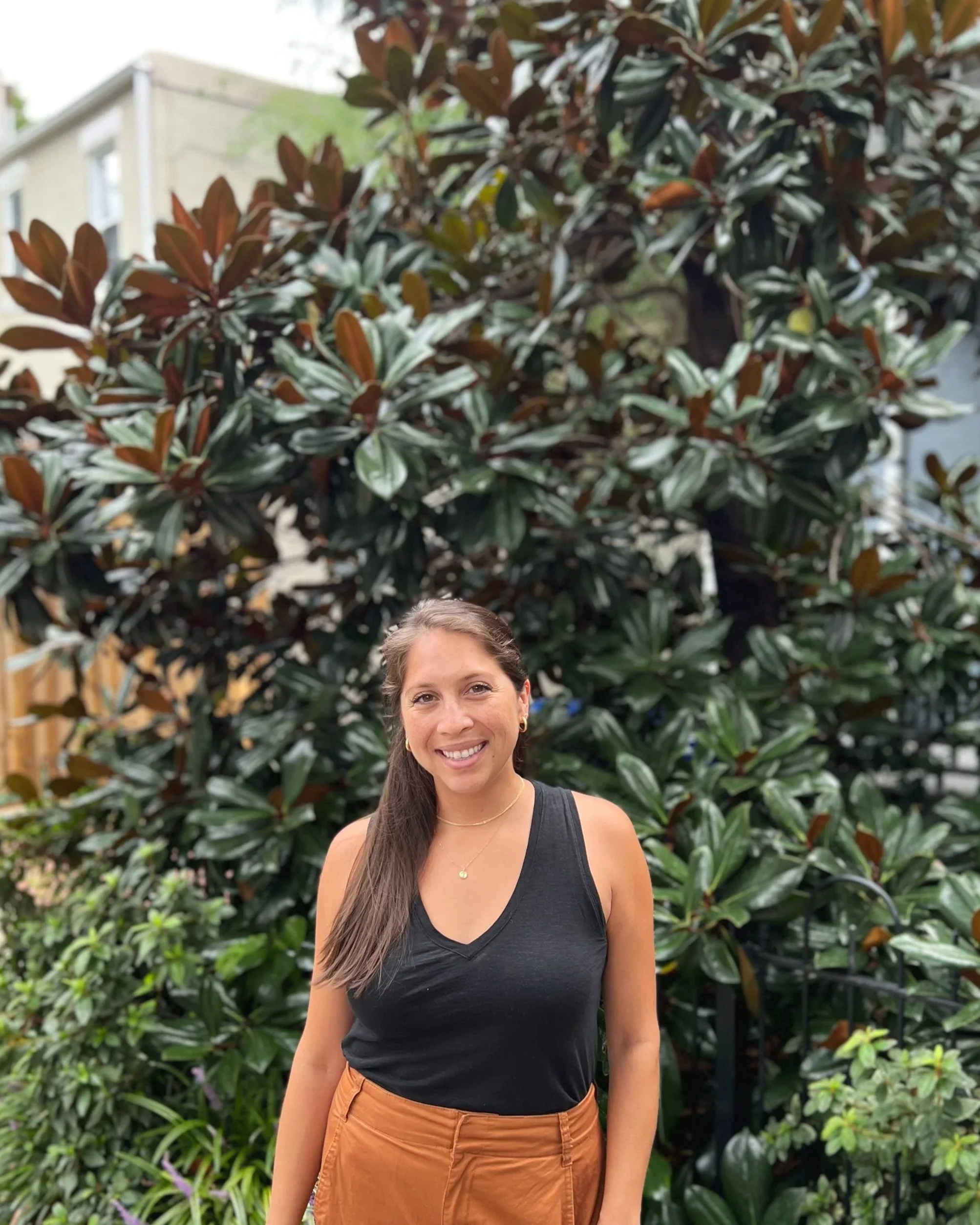 A woman with long hair smiling in front of a large leafy bush with dark green and reddish leaves.