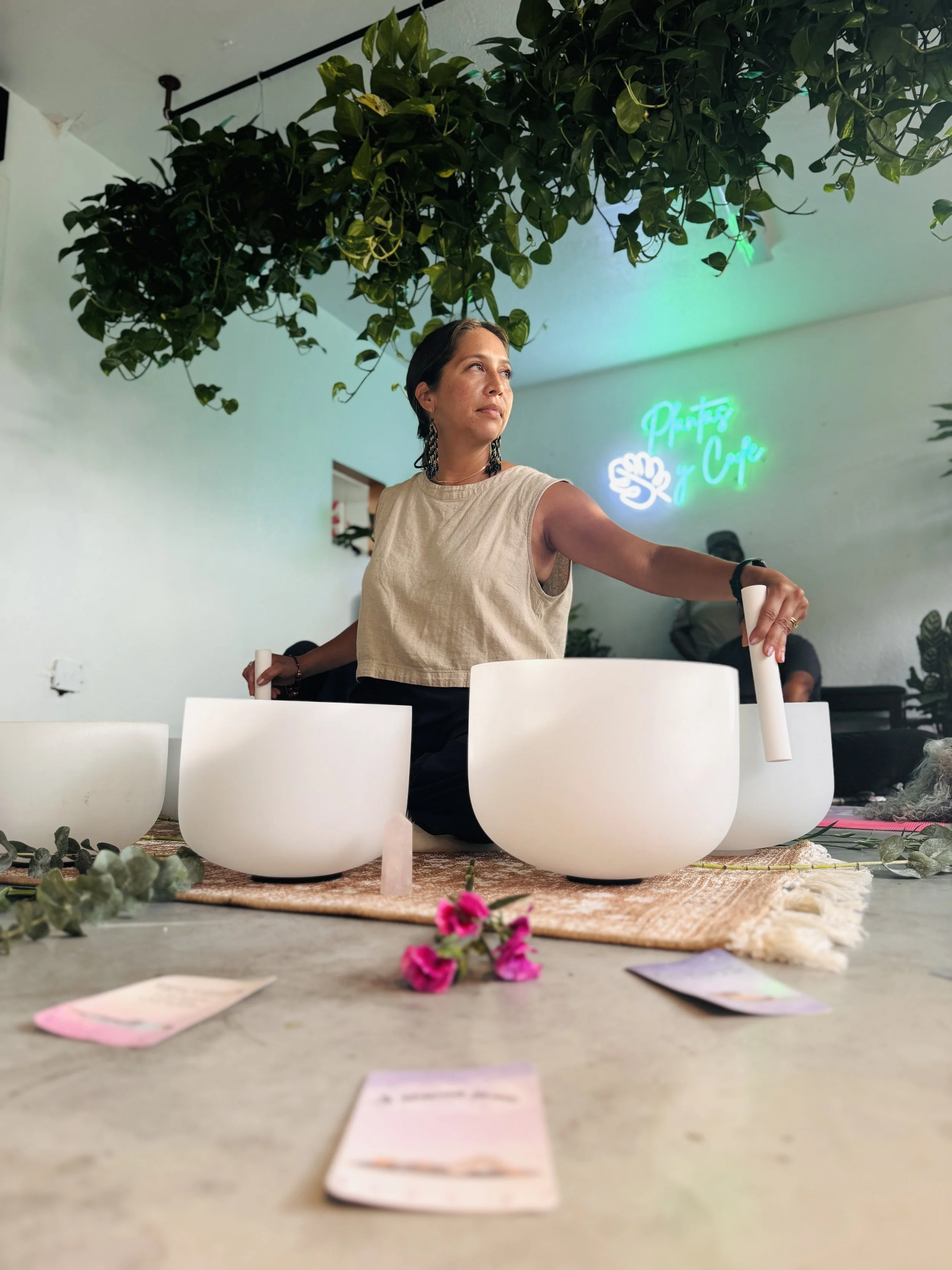 A woman with short dark hair and earrings participating in a sound healin session in a brightly lit room with a hanging plant, a neon sign on the wall, and white bowls on a woven mat on the floor decorated with pink flowers and small pieces of paper.