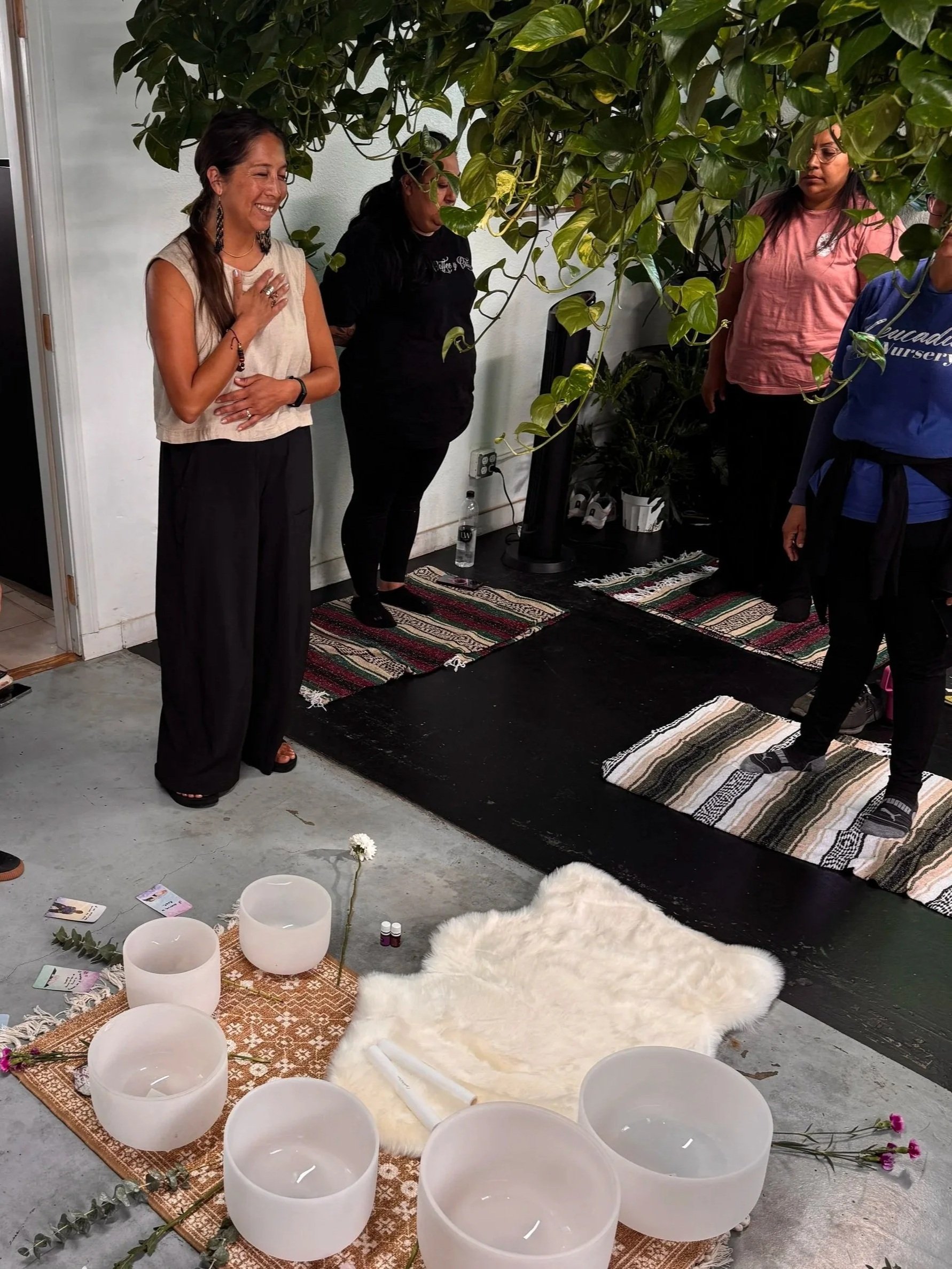 A group of women in a room with a large leafy plant, participating in a spiritual or meditation session. They are standing on small rugs, with singing bowls, flowers, and candles arranged on the floor in front of them.