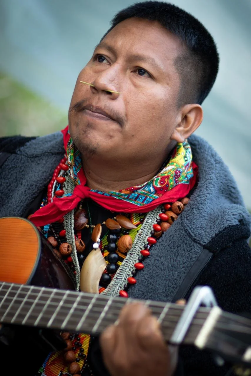 A man with short black hair and wearing colorful beaded necklaces and a vibrant scarf, playing an acoustic guitar.