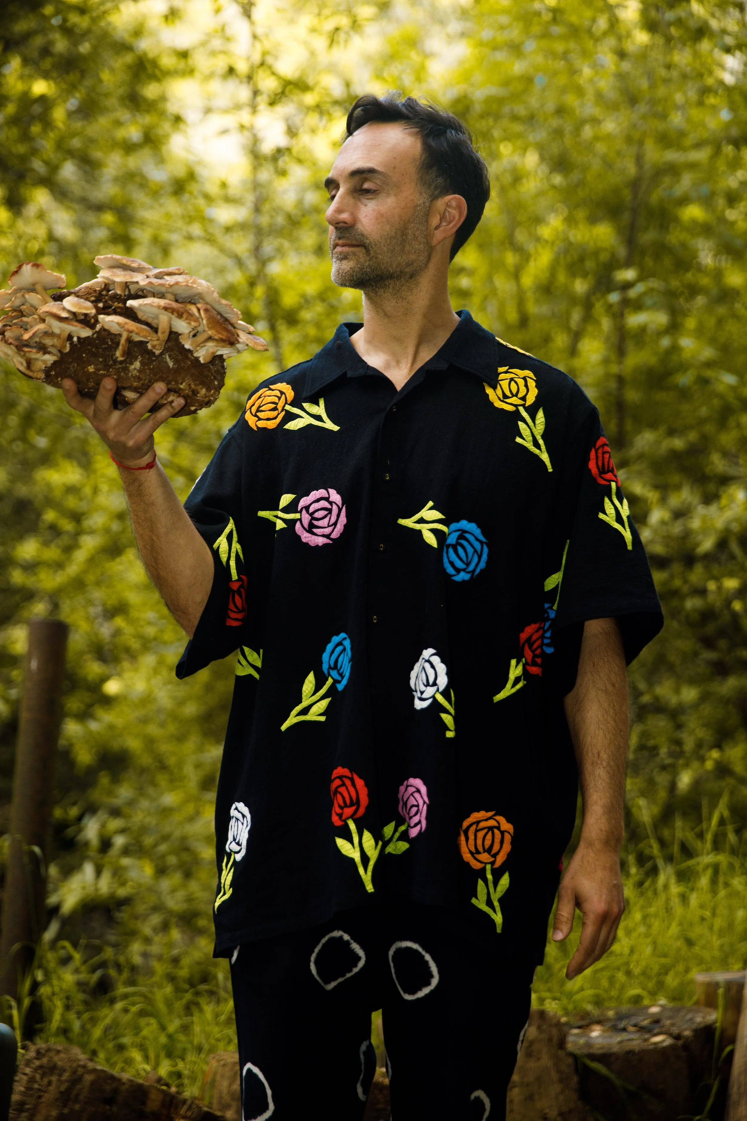 A man in an black shirt with roses deisgned holding a mushroom in his right hand