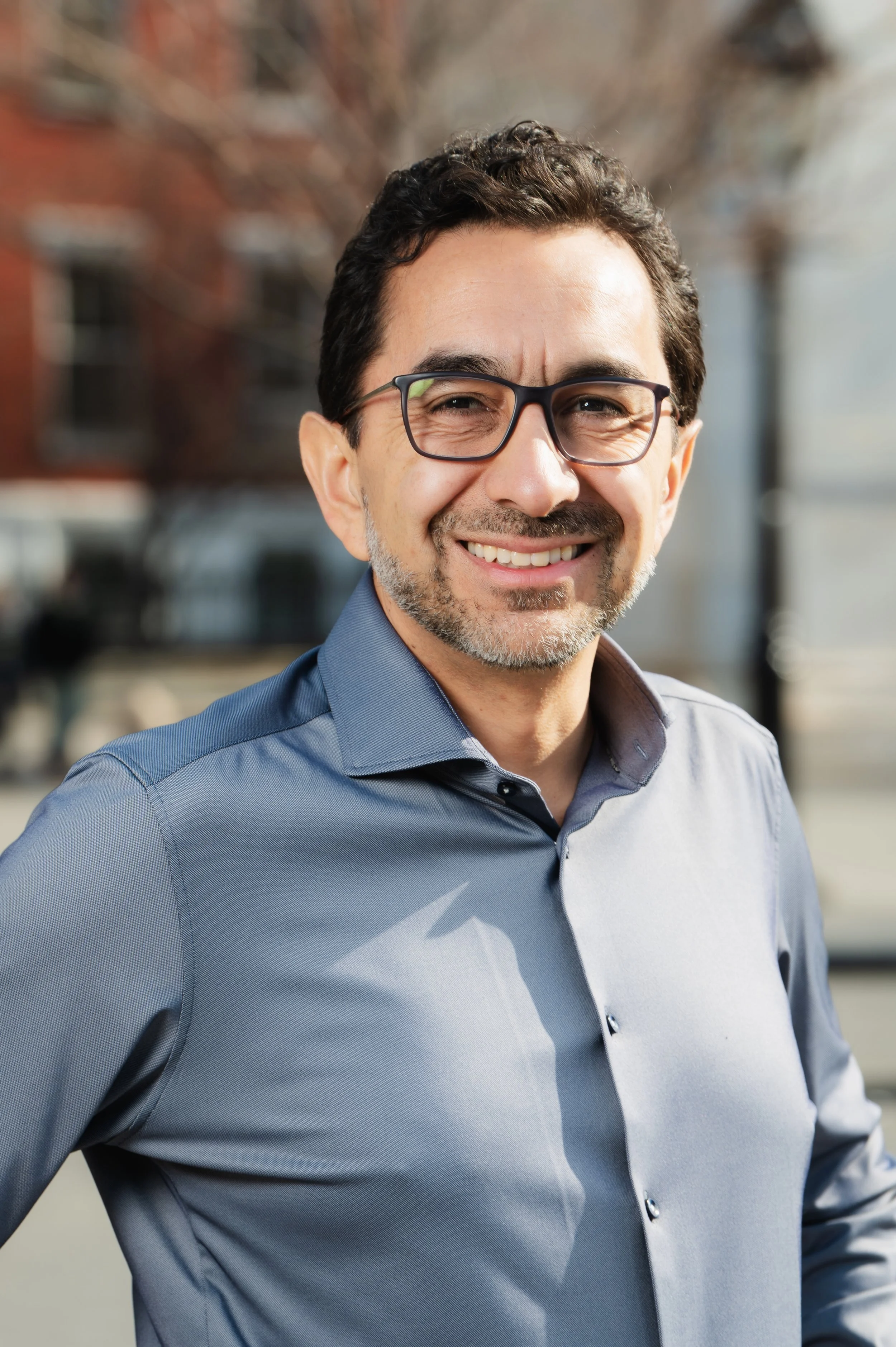 Portrait of a smiling man with glasses, wearing a gray button-up shirt, standing outdoors with blurred buildings and trees in the background.