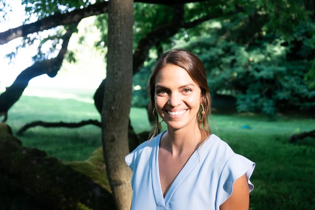 A woman with brown hair smiling outdoors, wearing a light blue top and gold earrings, standing near trees and greenery.