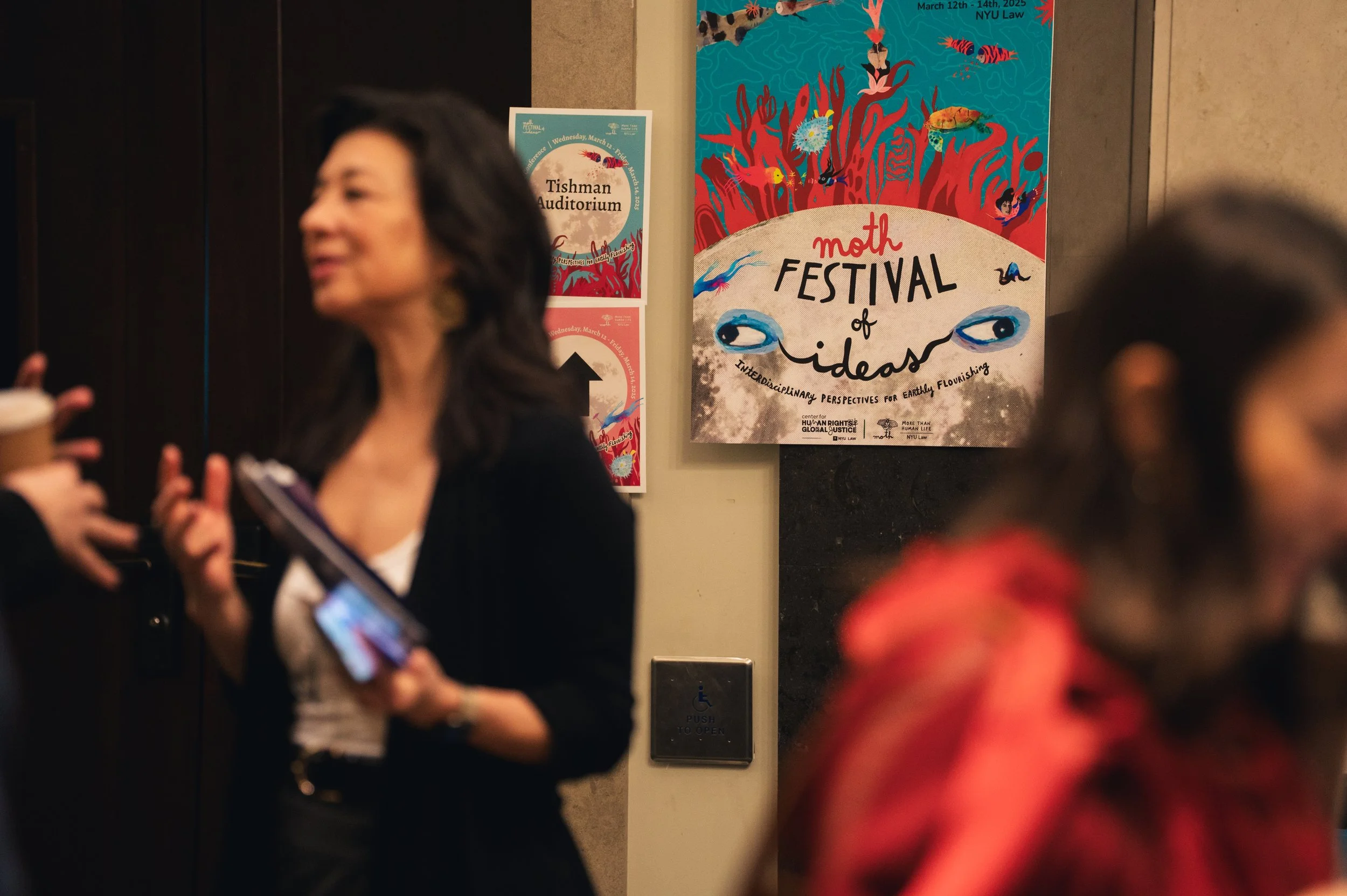 People attending the Moth Festival of Ideas with colorful posters on the wall, one of which reads "Moth Festival of Ideas" and features illustrations of eyes and underwater sea life.