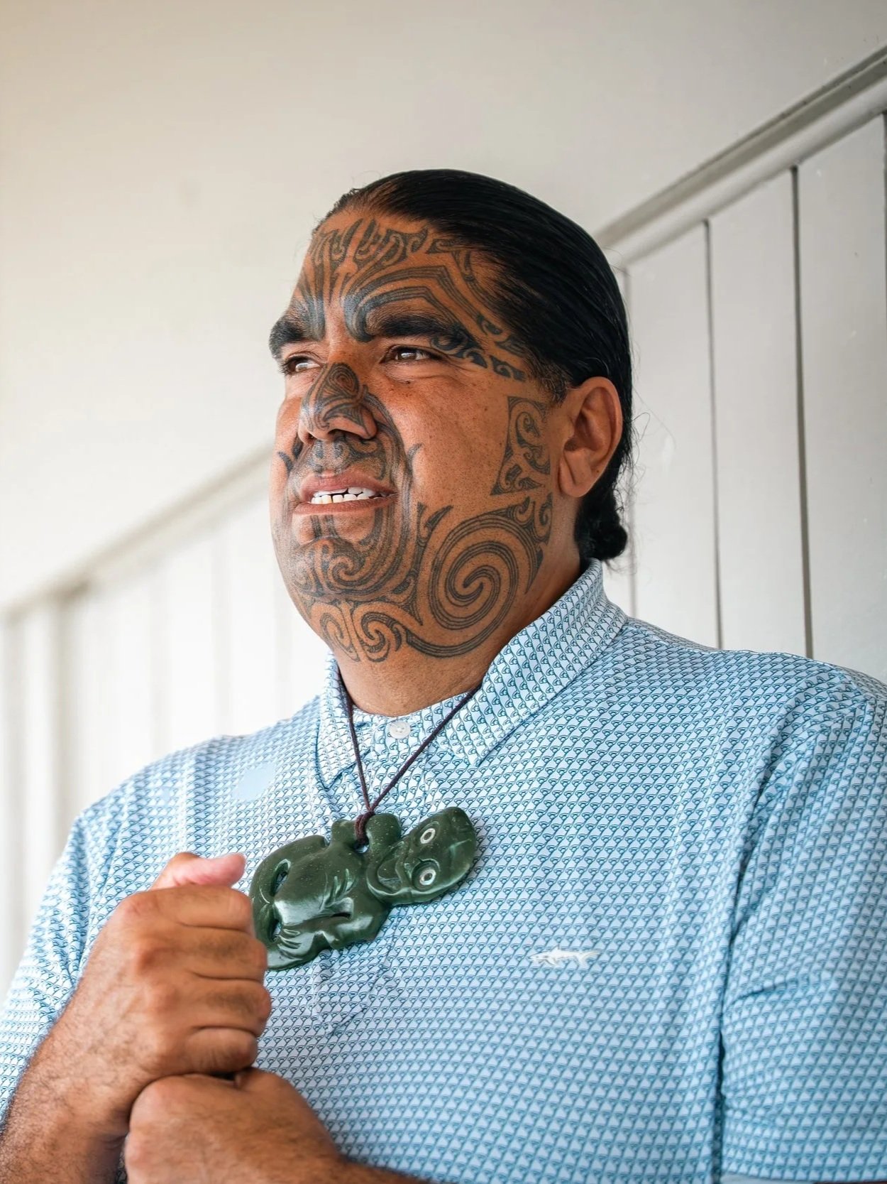 Man with traditional Maori tattoos on face and neck, wearing a blue patterned shirt and green jade necklace, standing indoors near white paneled wall.