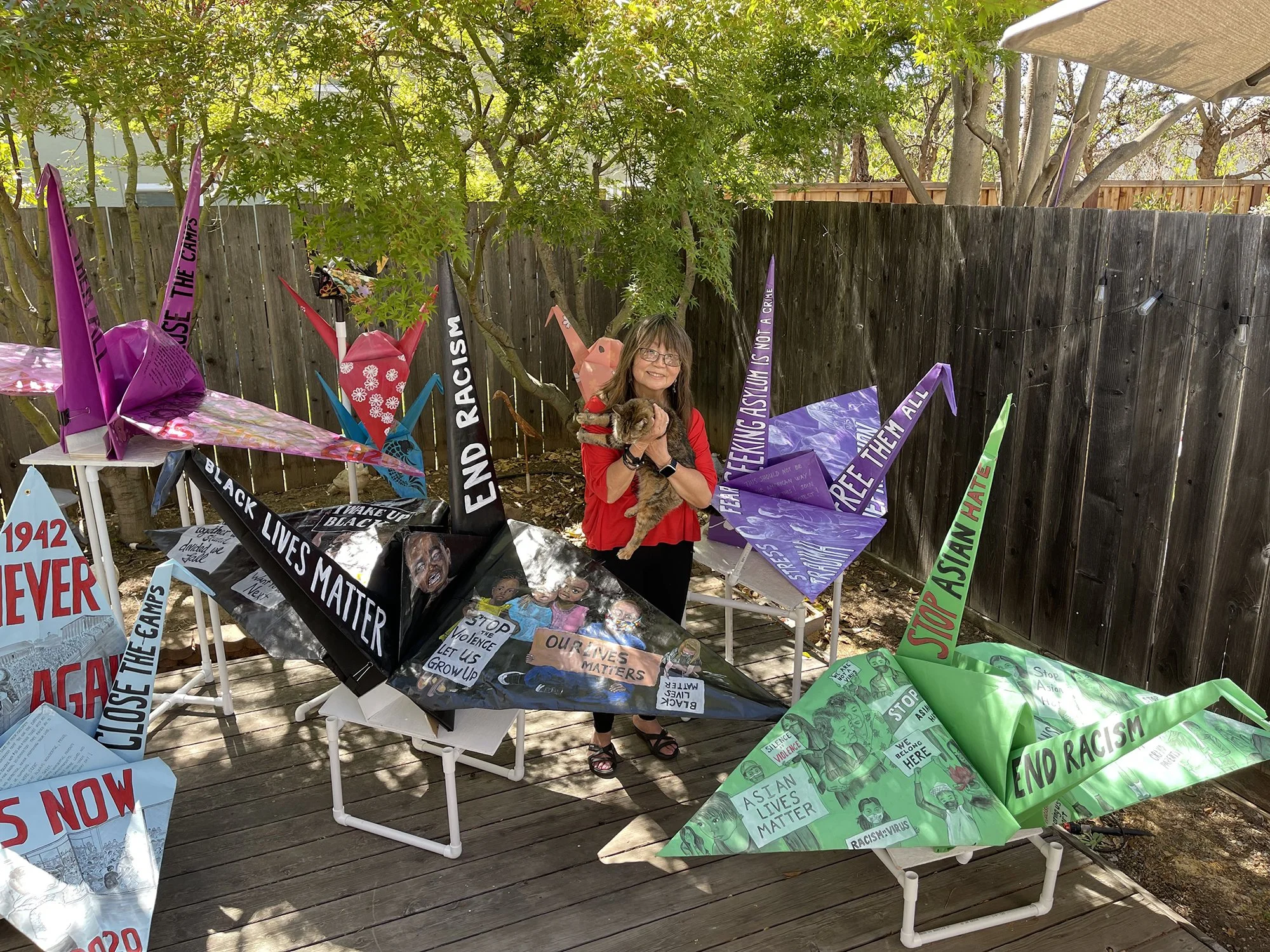 A person standing on a wooden deck surrounded by large origami cranes. Each crane is decorated with social justice messages and artwork, including phrases like "End Racism," "Black Lives Matter," and "Stop Asian Hate." The person is holding a dog and is in front of a wooden fence with trees in the background.