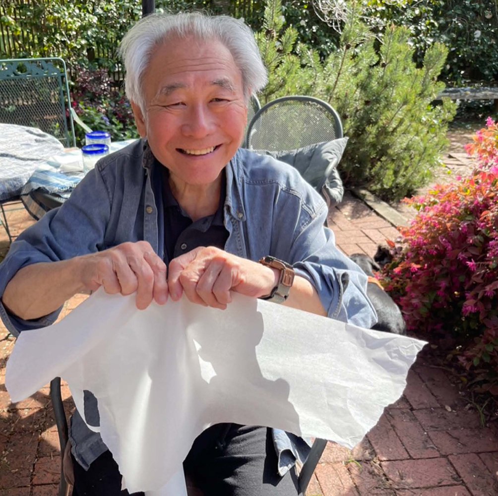 Smiling person seated outside, holding a sheet of white paper, surrounded by greenery.
