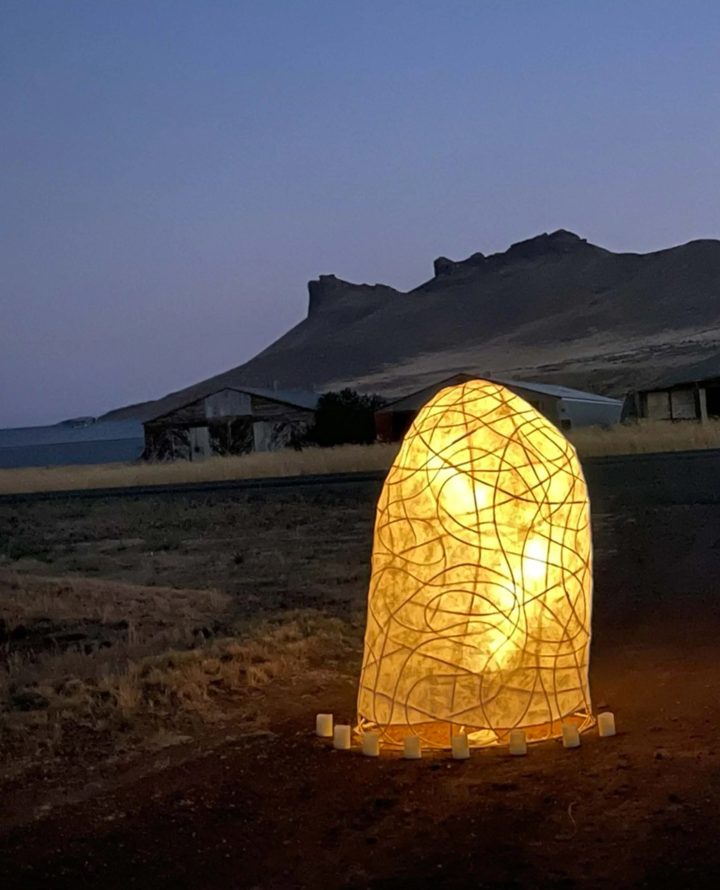 Glowing art installation resembling a lantern on a hilltop during twilight, with mountainous landscape in the background.