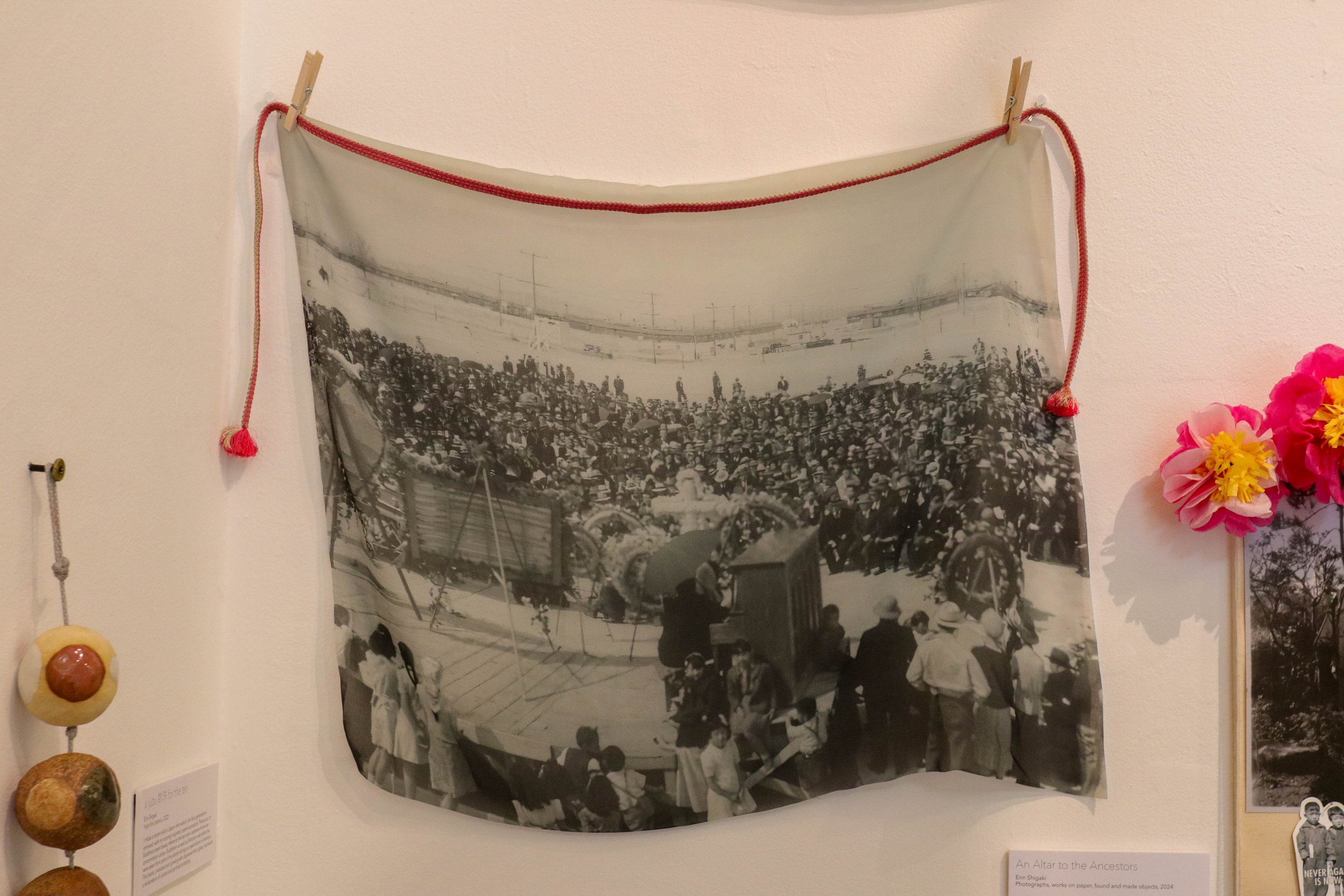 Vintage black and white crowd photograph on cloth, hanging with red rope and clothespins, accompanied by paper flowers and other art objects.