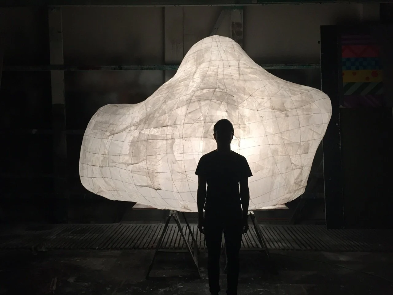 Silhouette of a person standing in front of a large, glowing, abstract paper lantern sculpture in a dimly lit room.