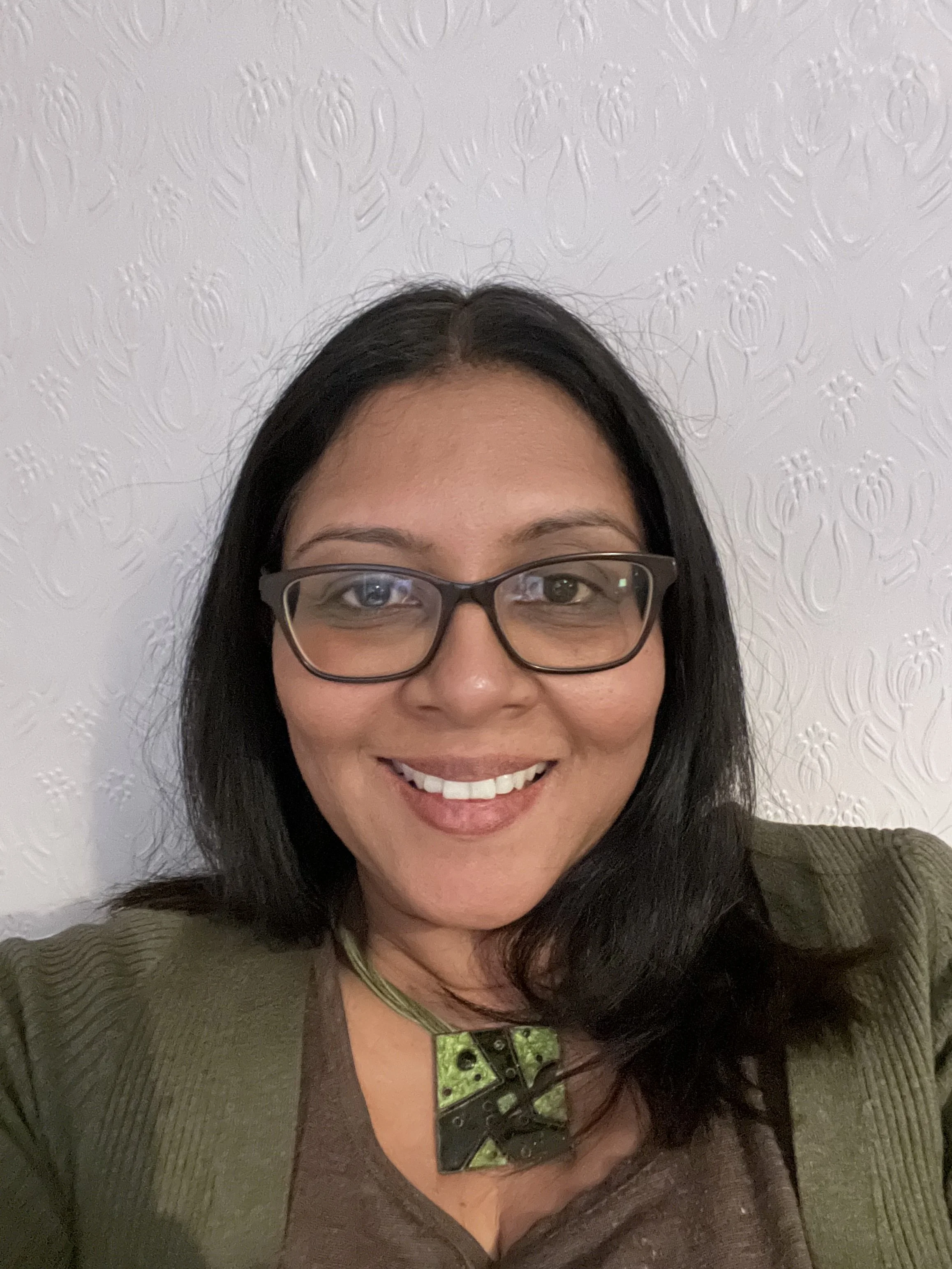 A woman with shoulder-length dark hair, wearing glasses, a brown shirt, a green jacket, and a green necklace, smiling while taking a selfie in front of a textured white wall.