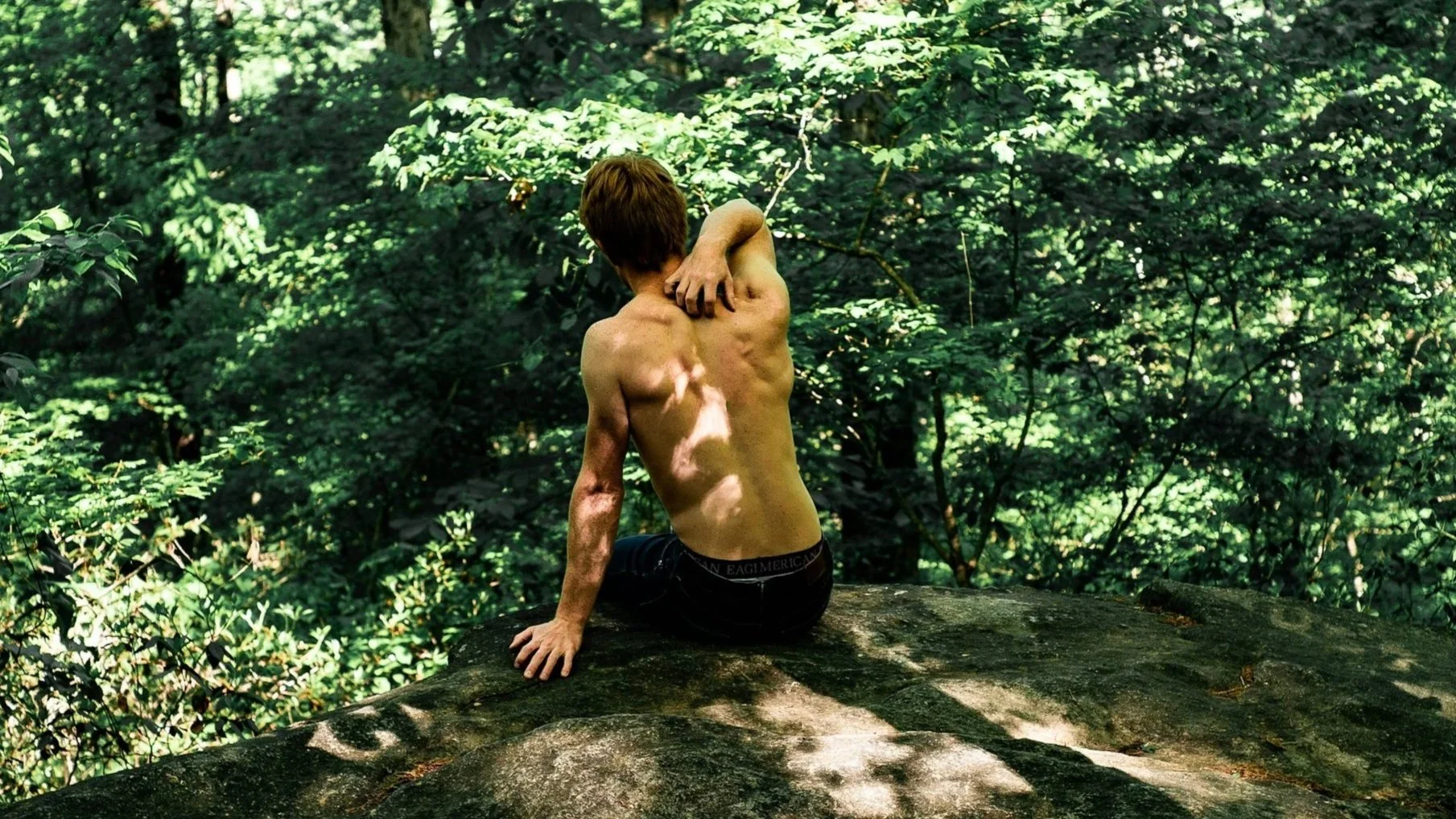 White man sitting on the edge of a rocky hill while leaning on his left side and holding his right upper trap with his right hand