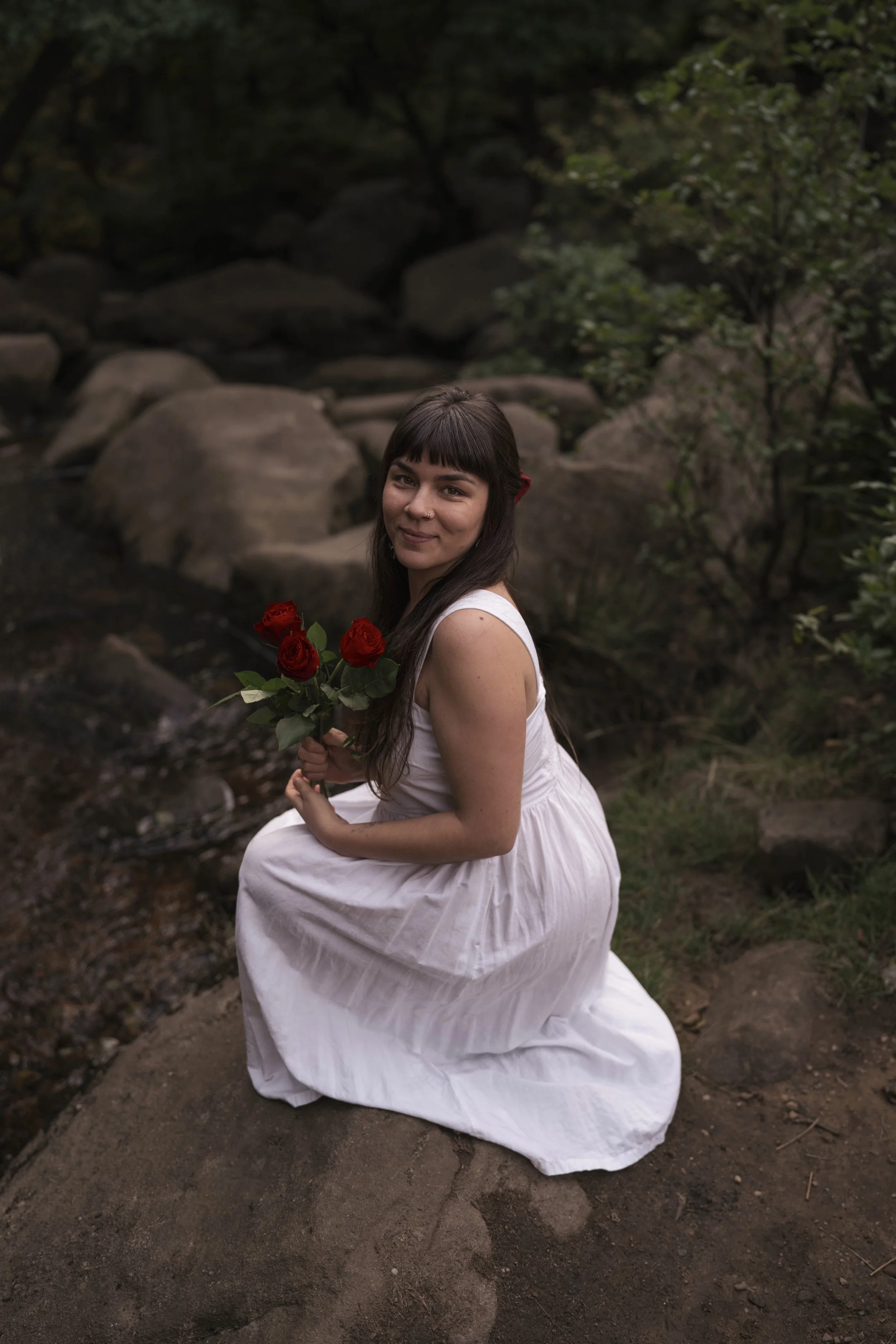 A woman with dark hair and bangs, wearing a white dress, is kneeling on one knee outdoors near rocks and greenery, holding a bouquet of red roses.
