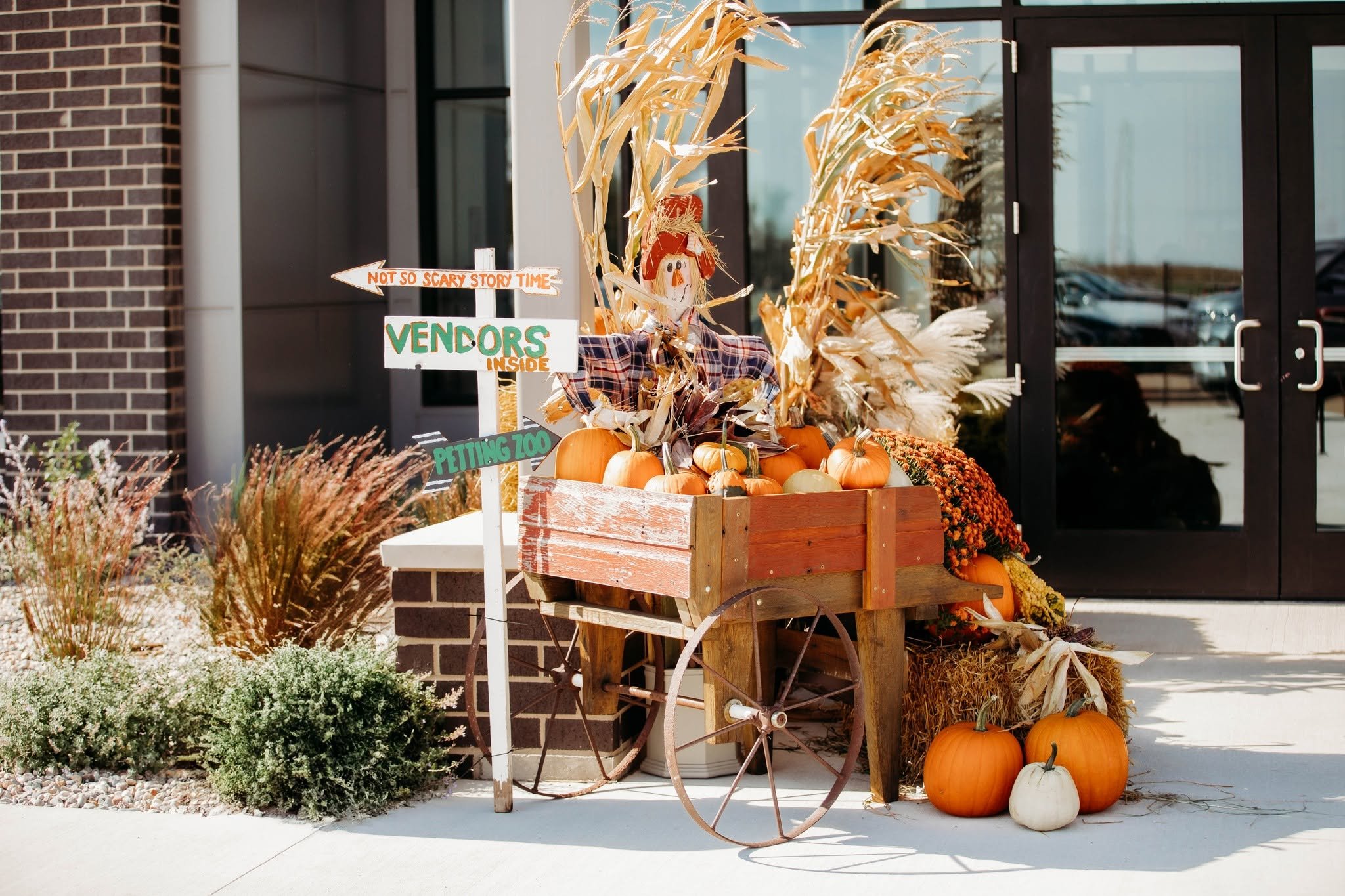 Orange and white pumpkins in a wooden cart outside a building decorated for autumn, with dried corn stalks and a scarecrow figure. Signage indicates activities inside and different event themes.