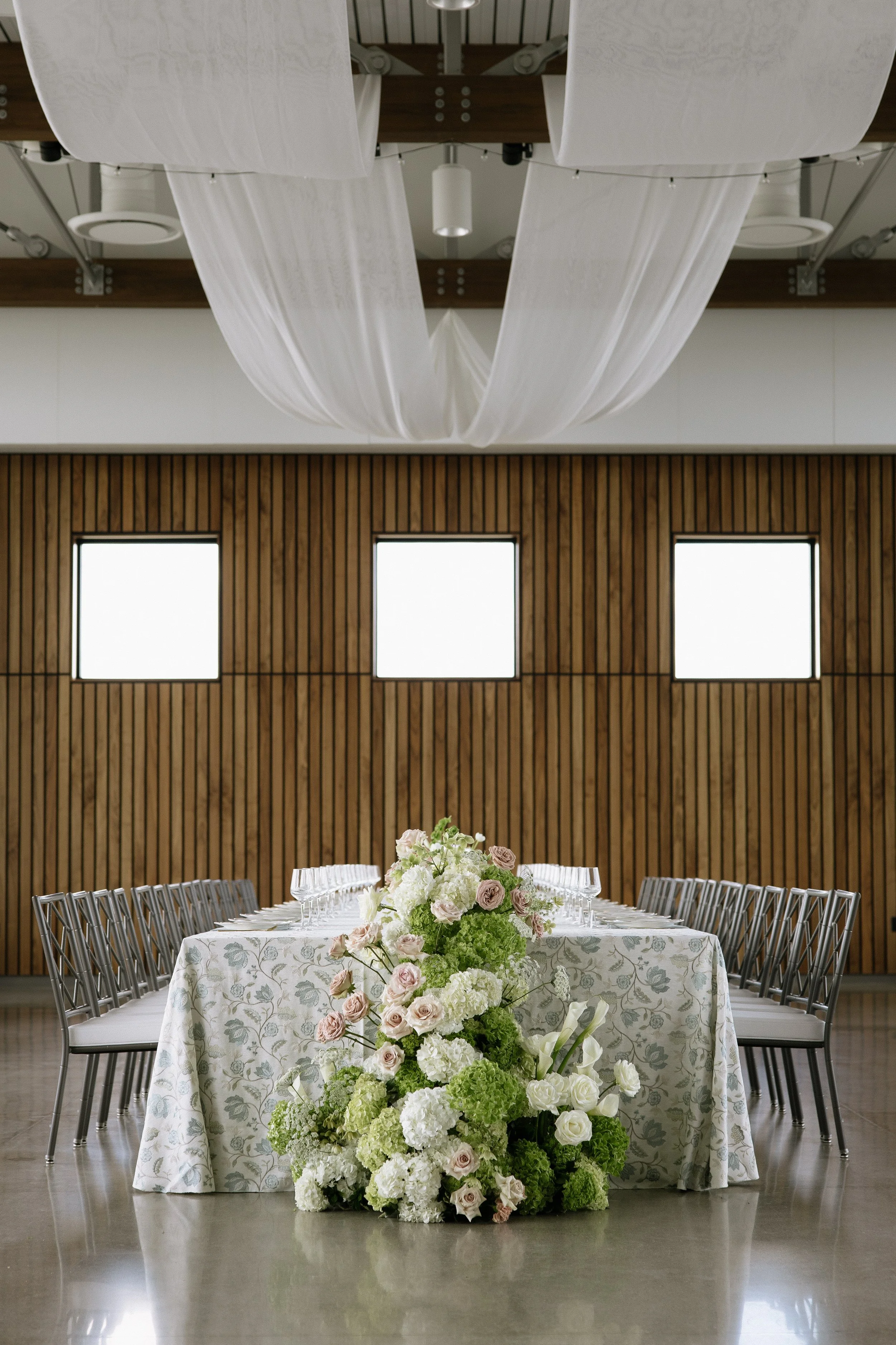 A long harvest wedding head table with a soft floral-patterned linen from Reverie Social set beneath white ceiling draping. A lush arrangement of white, blush, and green flowers cascades from Love Always Floral. Framed by modern silver chairs and a w