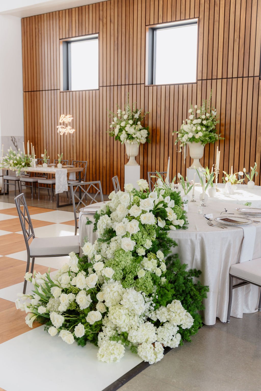 Elegant wedding reception setup with white floral arrangements, round tables with white tablecloths, and wooden wall backdrop with windows. Sign reading 'Better Together' is illuminated on the wall.