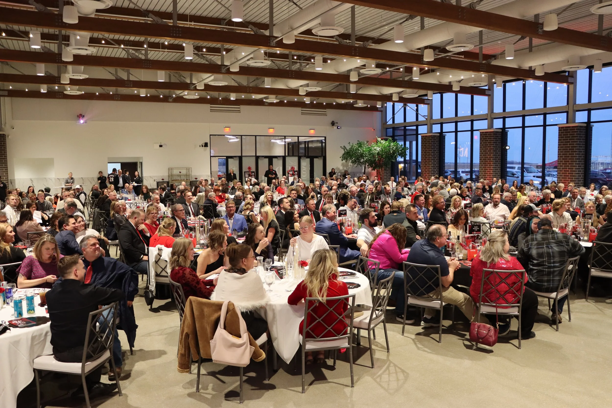 View of a large banquet hall filled with numerous guests seated at round tables, decorated for a formal event with red accents, with large floor-to-ceiling windows showing an evening view outside and the ceiling featuring industrial design with expos