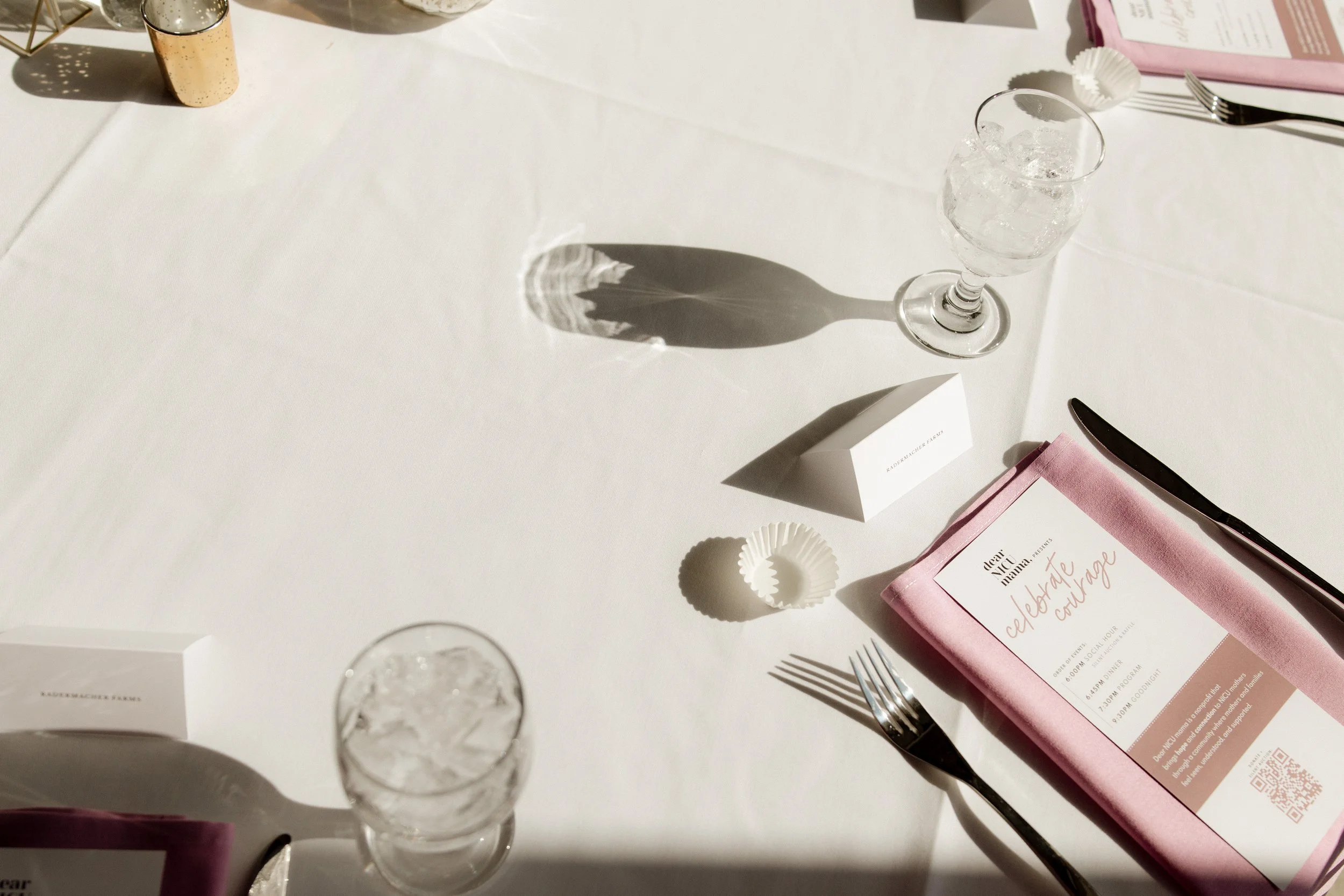 Table setting at an event with silverware, pink napkins, a glass of ice water, a pink booklet or program, and small decorative items, all illuminated by sunlight creating shadows.