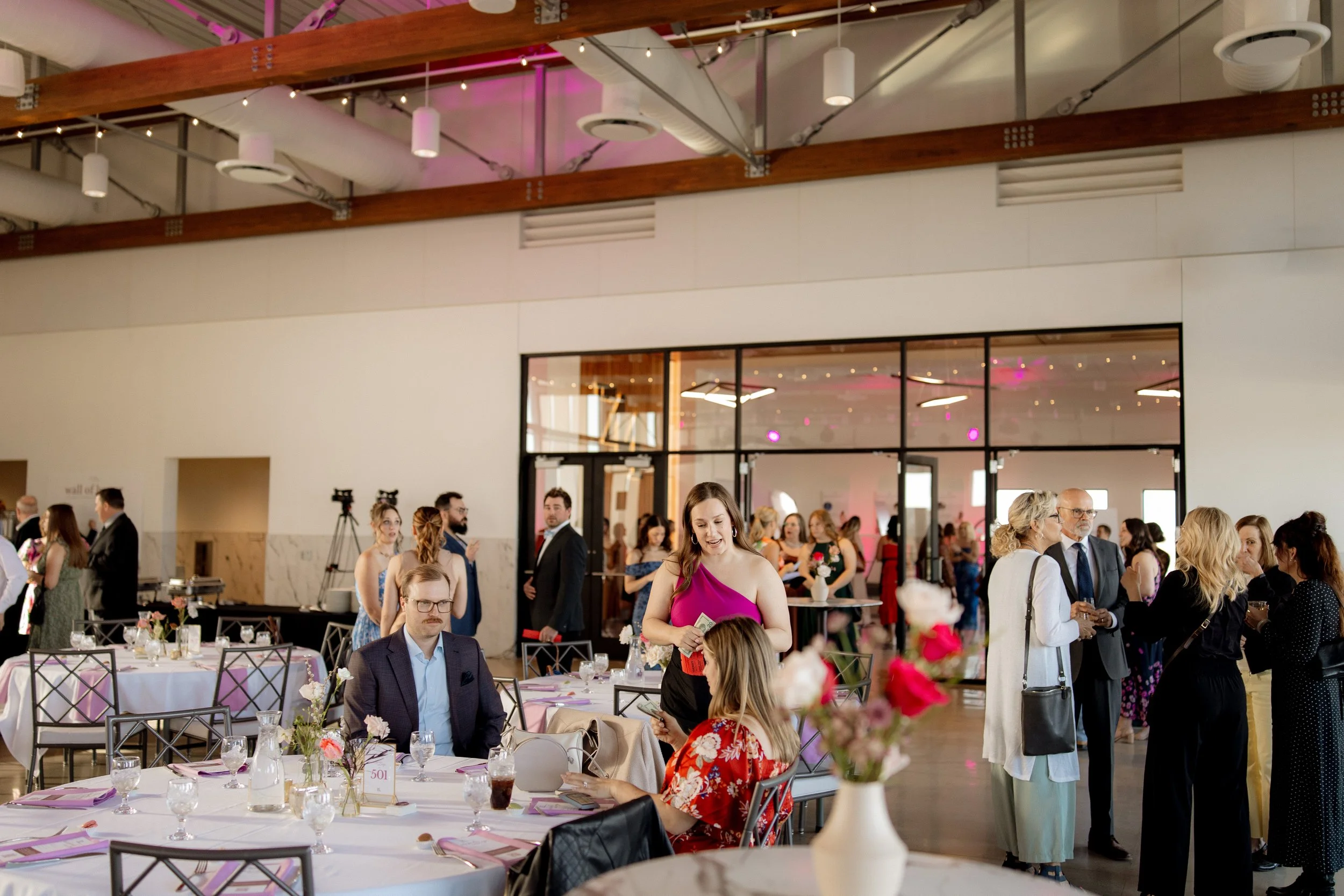 Guests gather at a wedding reception in a modern, spacious venue with high ceilings, exposed ductwork, and wooden beams. Some are standing, chatting and holding drinks, while others sit at tables decorated with floral centerpieces. The background sho