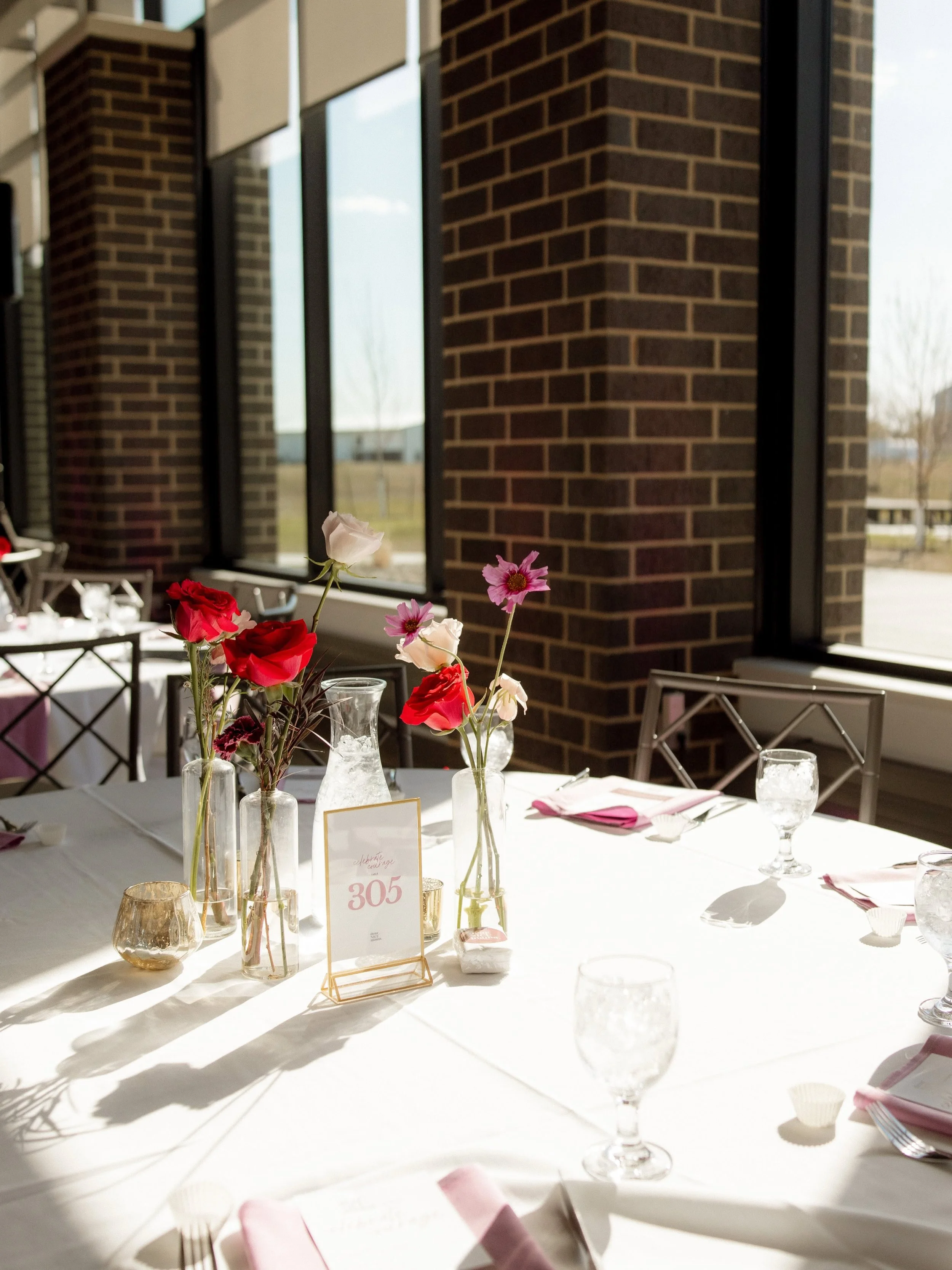 A decorated dining table set for an event inside a room with large windows and brick walls. The table has floral centerpieces with pink, red, and white flowers in vases, gold candles, pink napkins, and place settings with glassware and utensils, incl