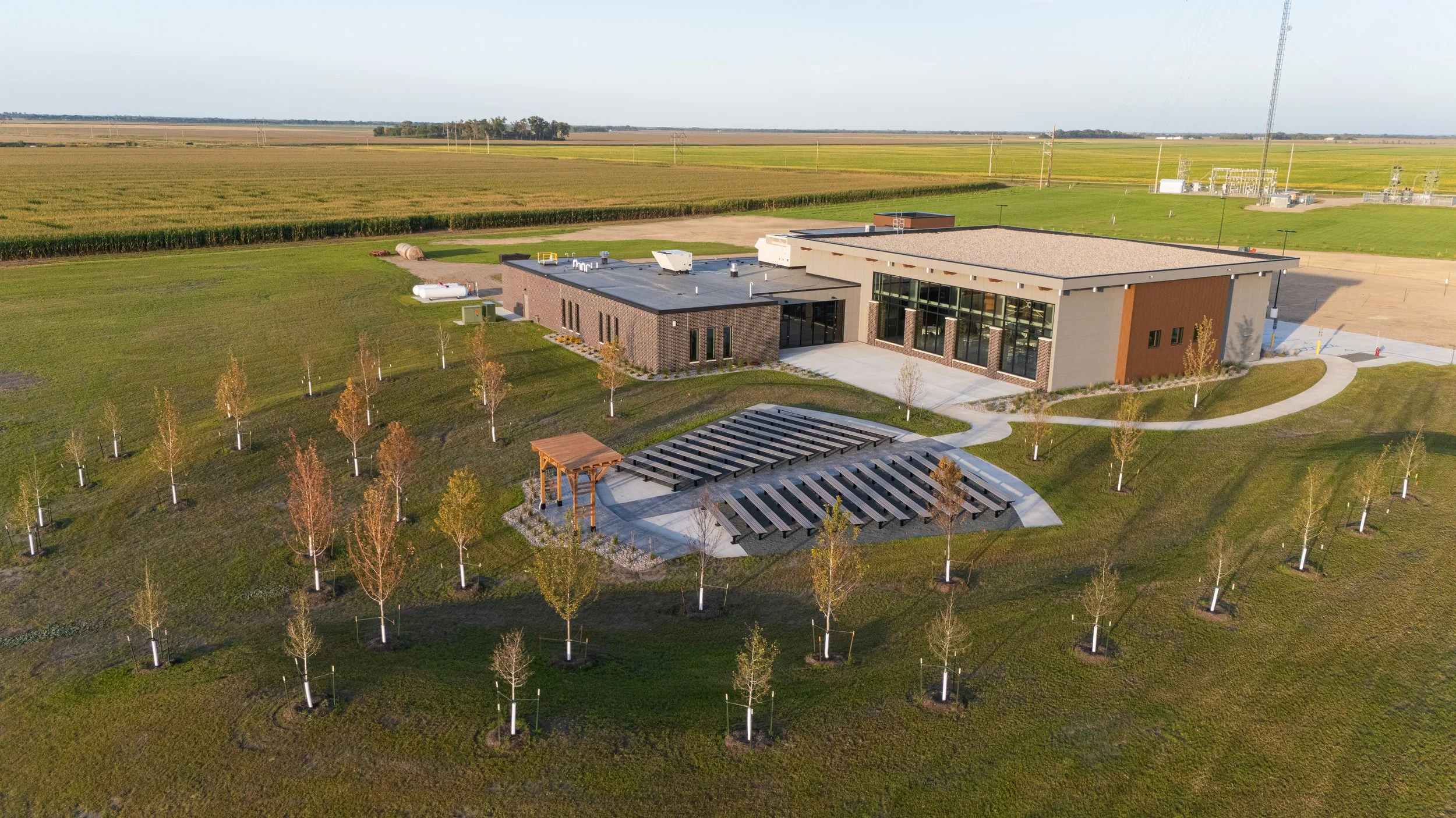 An aerial view of a modern building with surrounding landscaped area, trees, and solar panel structures. The building is situated in a rural area with fields and power lines in the background.