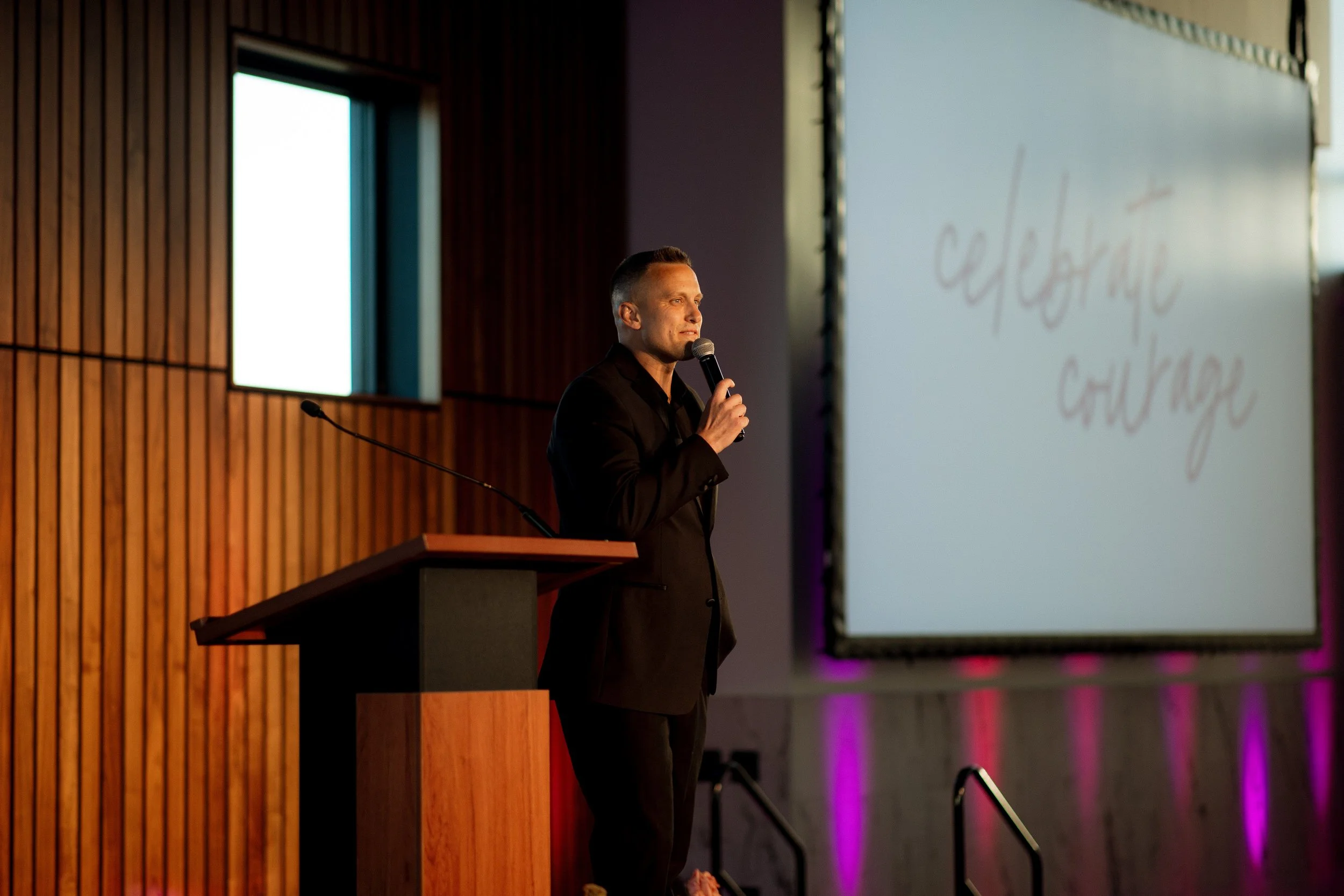 A man in a black suit holding a microphone speaks at a podium in a conference room with wood paneling and a large screen displaying the words 'celebrate Chyzok' in cursive.