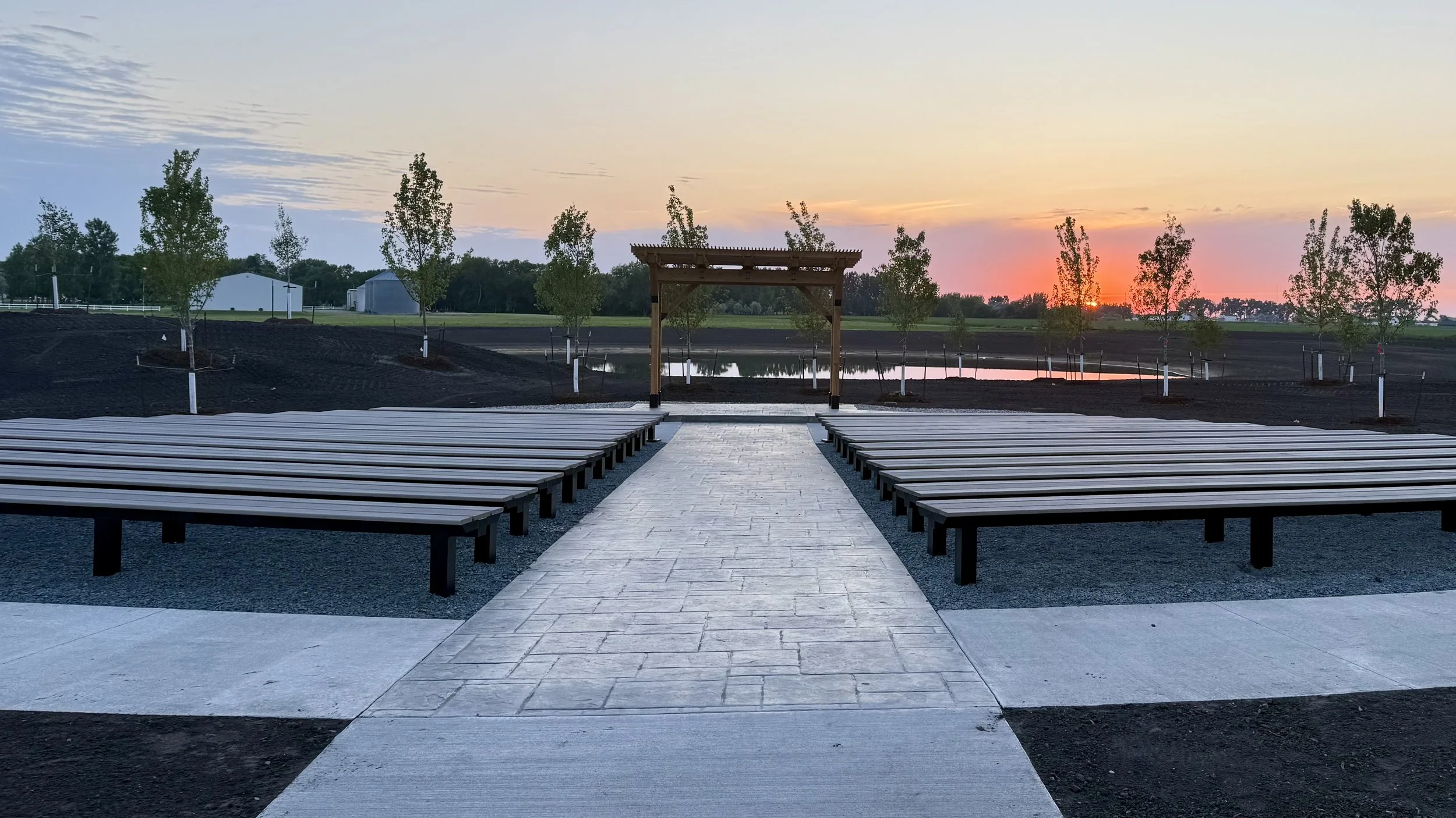 Sunset over a landscaped park with benches, a pathway, and a wooden entrance gate, overlooking a pond with trees in the background.