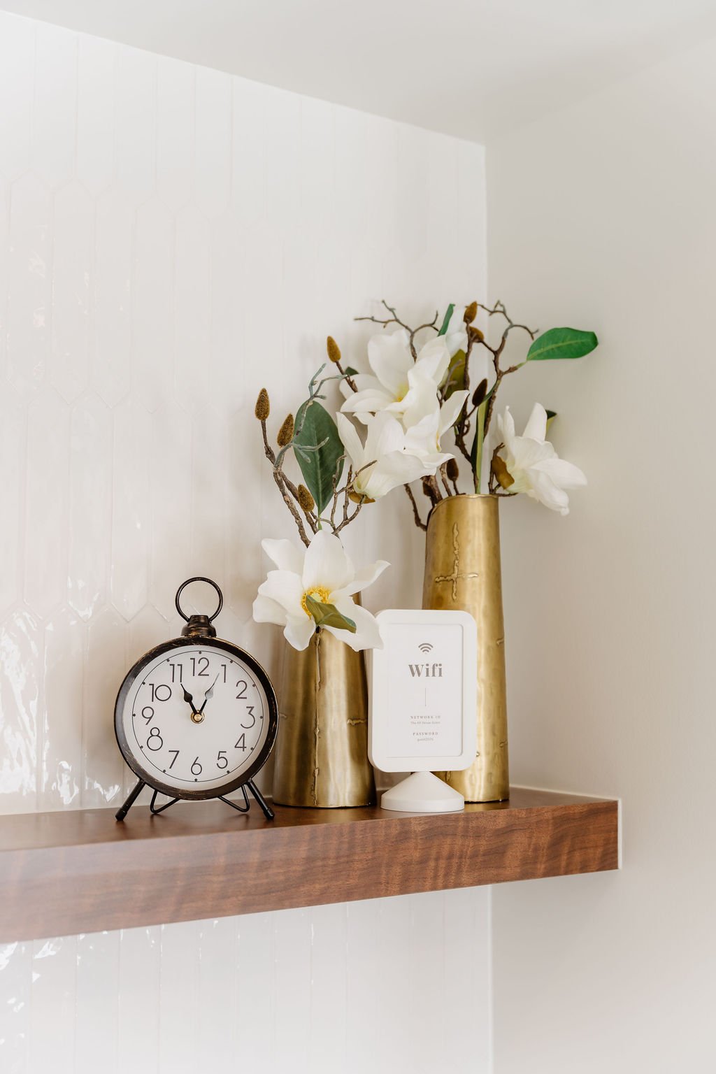 Decorative wall shelf with a black clock, two gold vases with white flowers and greenery, and a white Wi-Fi sign.