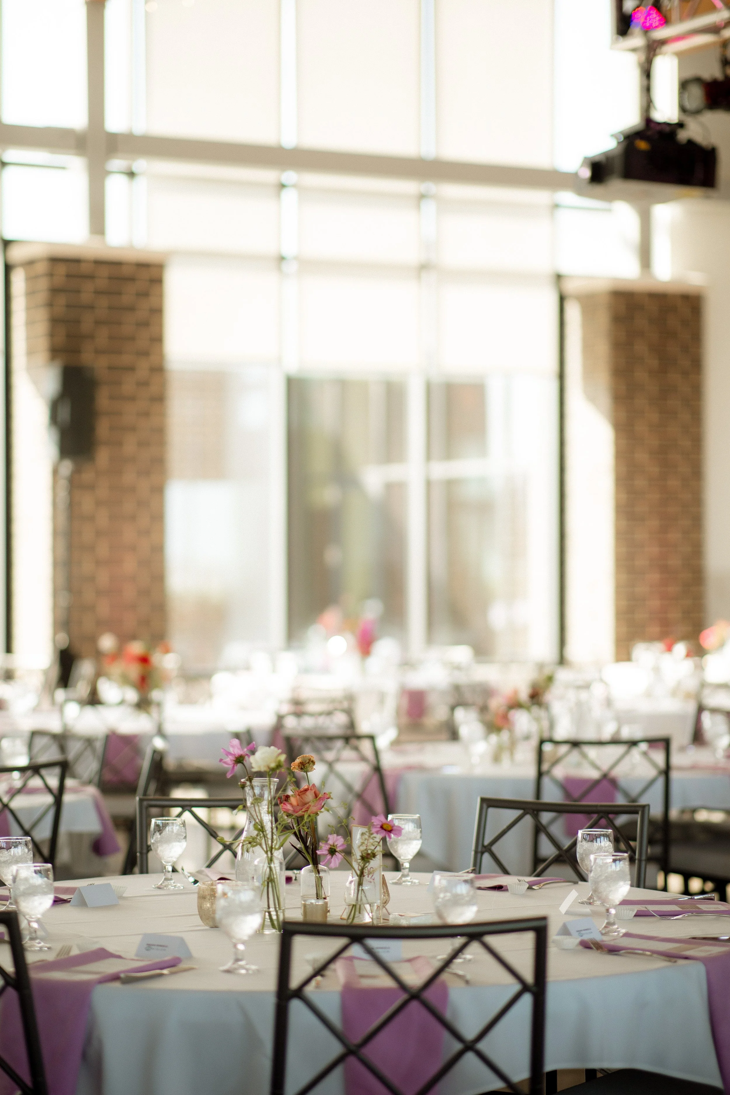 Interior of a banquet hall with round tables decorated with floral centerpieces, wine glasses, and pink napkins, set for a formal event, with large windows in the background.