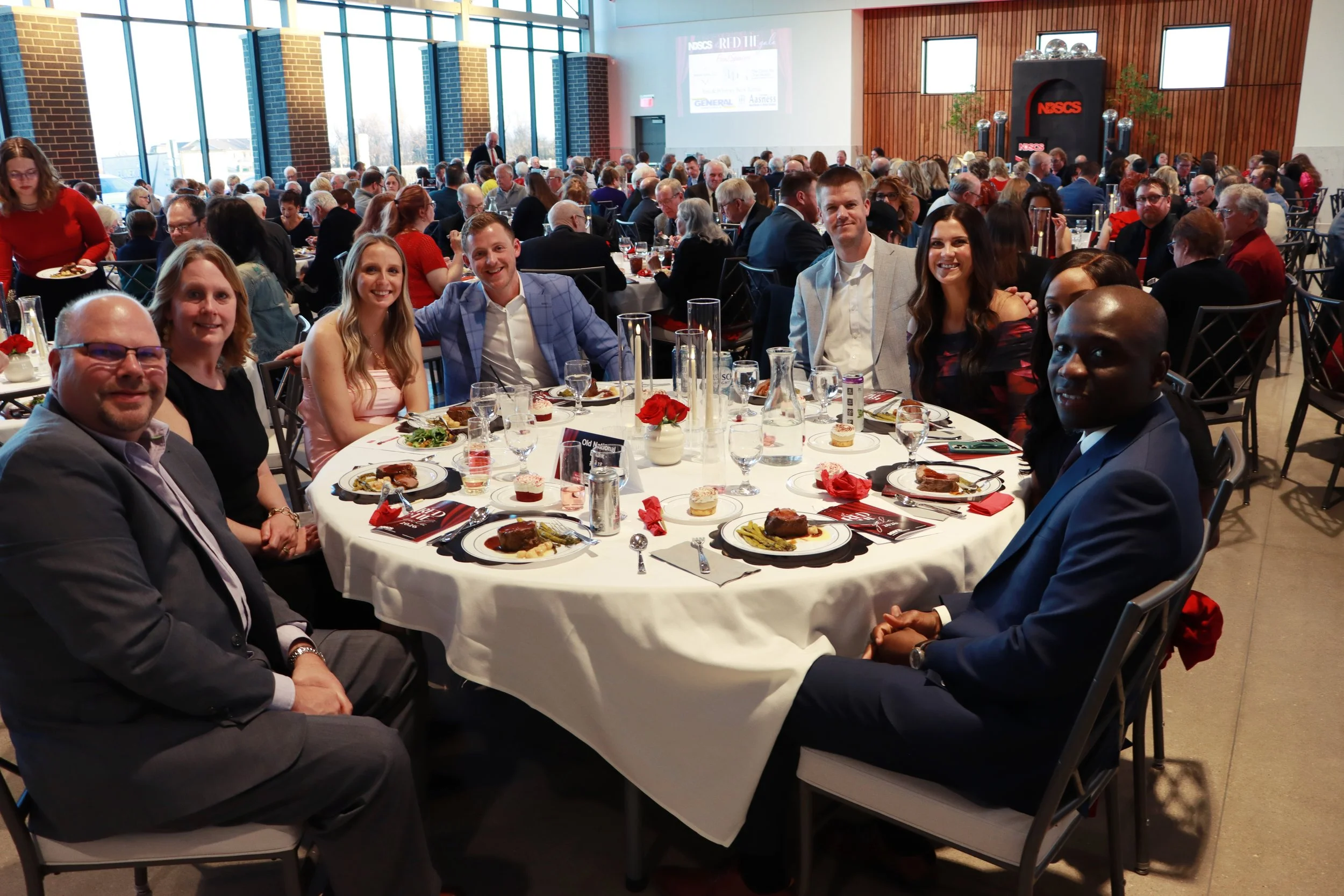 A large gathering at a formal event or banquet inside a spacious hall with floor-to-ceiling windows. The tables are decorated with red flowers and full place settings. Attendees are dressed in formal and semi-formal attire, and the atmosphere appears