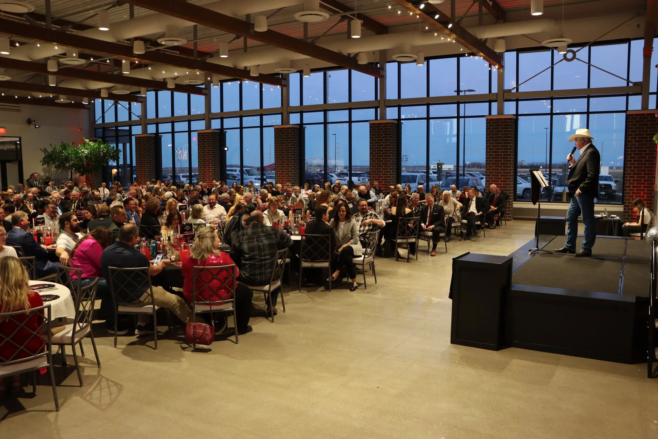 Large indoor event with many seated guests, a person in western attire speaking into a microphone on stage, and large windows showing an outdoor parking lot at dusk.