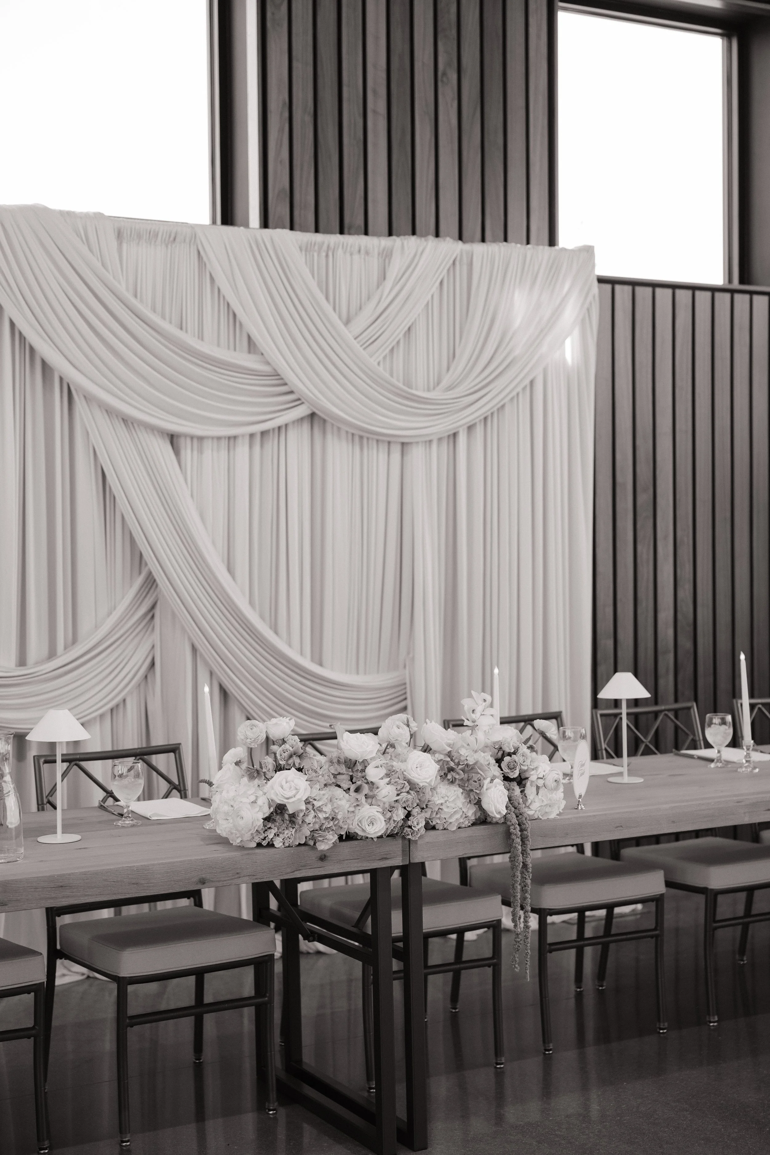 A black-and-white view of a long wooden wedding head table decorated with a lush floral arrangement, taper candles, and small table lamps. Layered white draping creates a dramatic backdrop against the venue’s tall windows and dark wood-paneled walls.