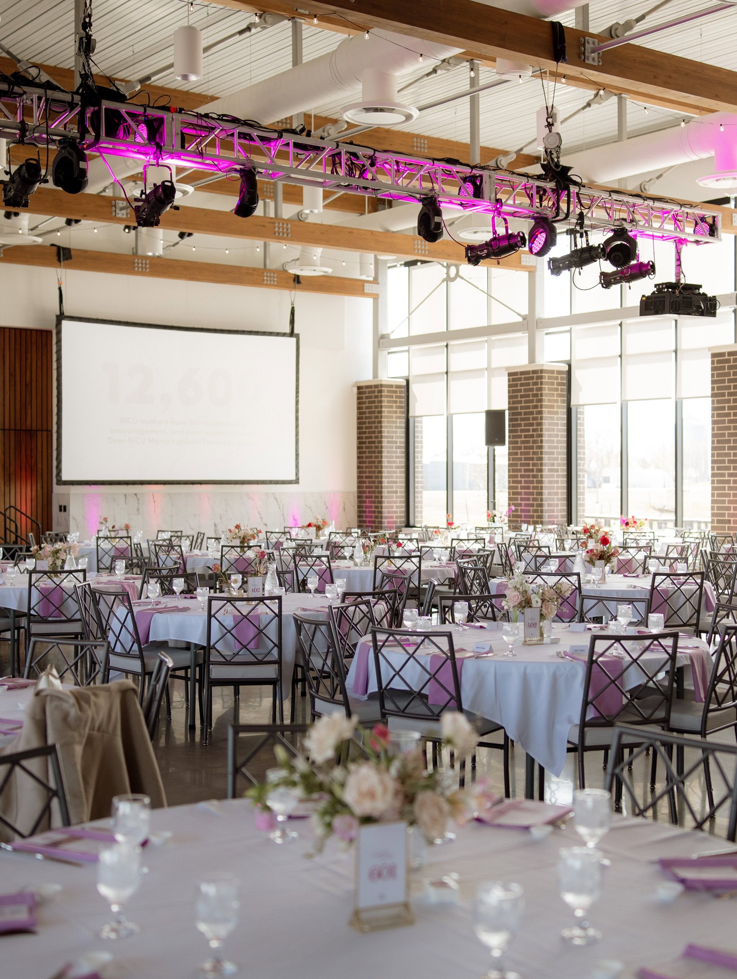 Elegant indoor wedding reception setup with decorated tables, floral arrangements, white drapes hanging from the ceiling, and a checkered dance floor in a modern venue.