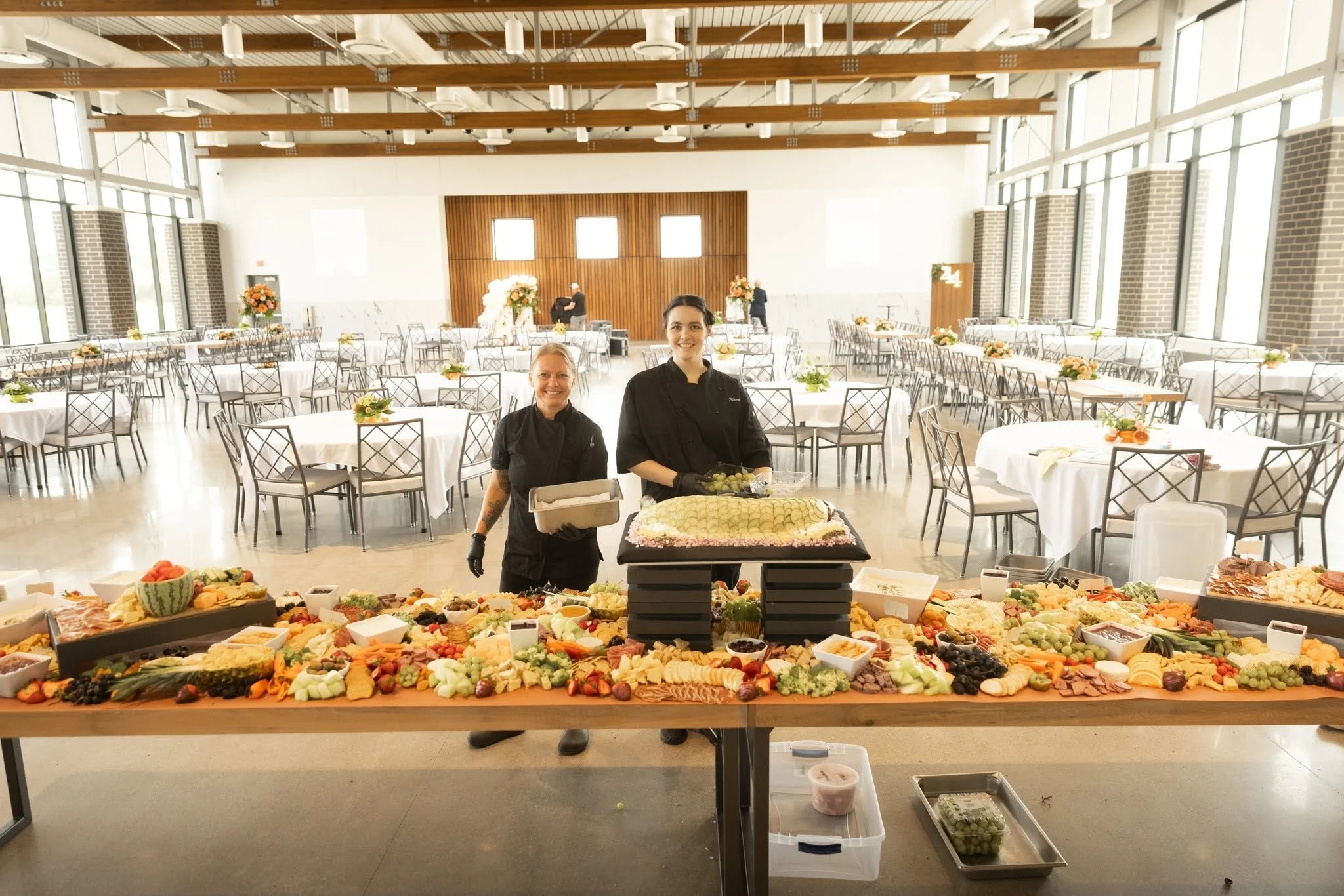 Two female chefs stand behind a large buffet table filled with various fruits, vegetables, and appetizers, in a spacious event hall with round tables adorned with flower arrangements.