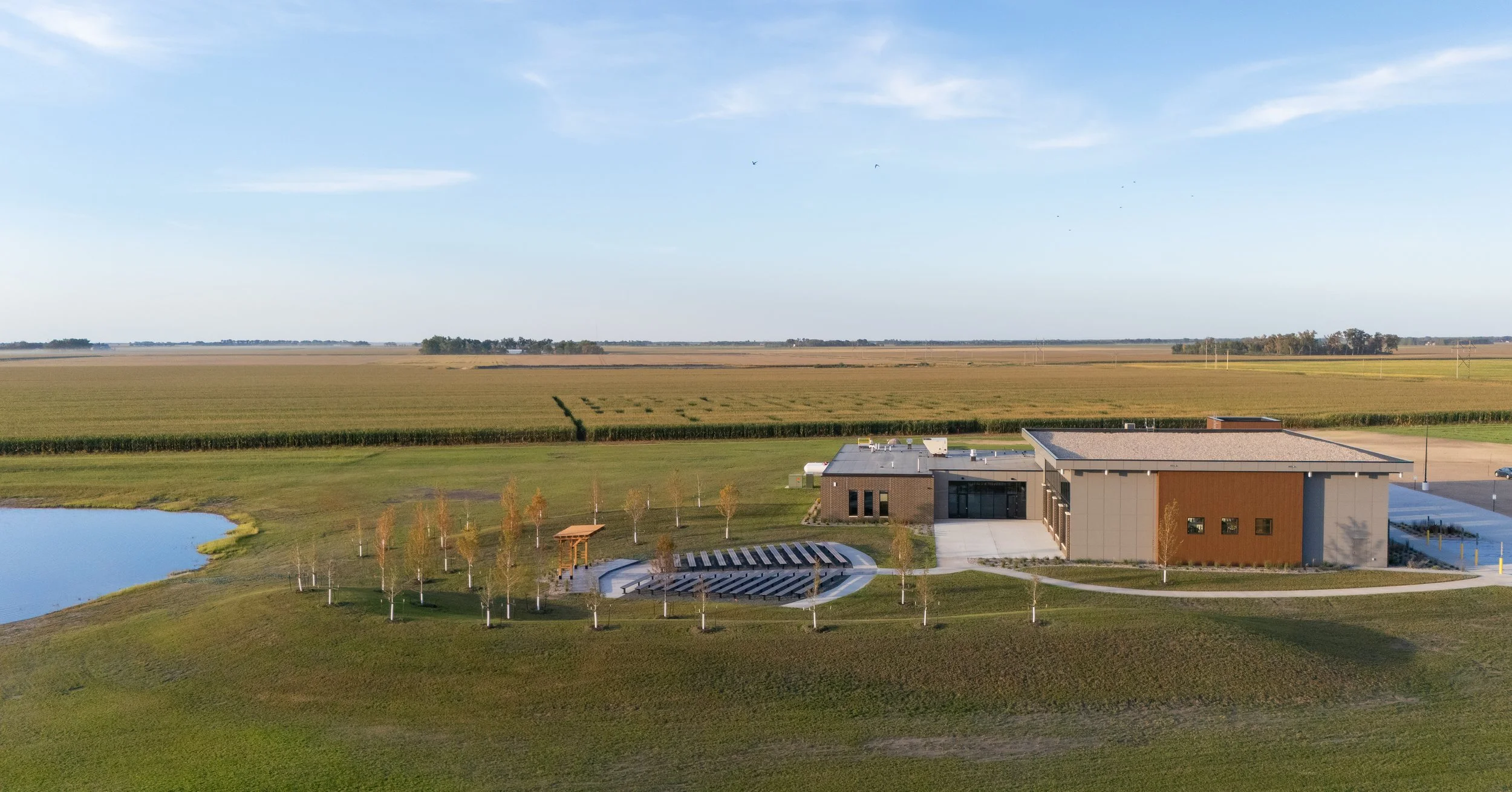 An aerial view of a modern building with outdoor seating and a small pond, surrounded by green grass and trees, with farmland and open fields in the background under a clear blue sky.