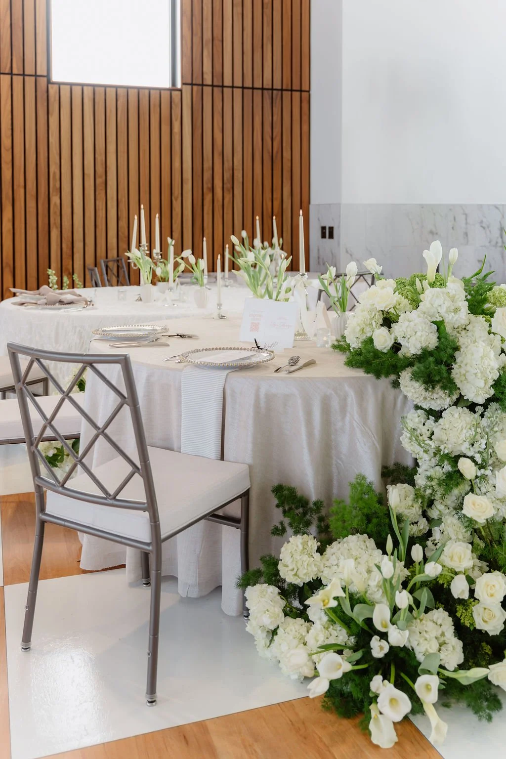 A beautifully decorated banquet table with white floral arrangements, candles, and elegant tableware in a modern, minimalistic room with wooden paneling and white marble walls.