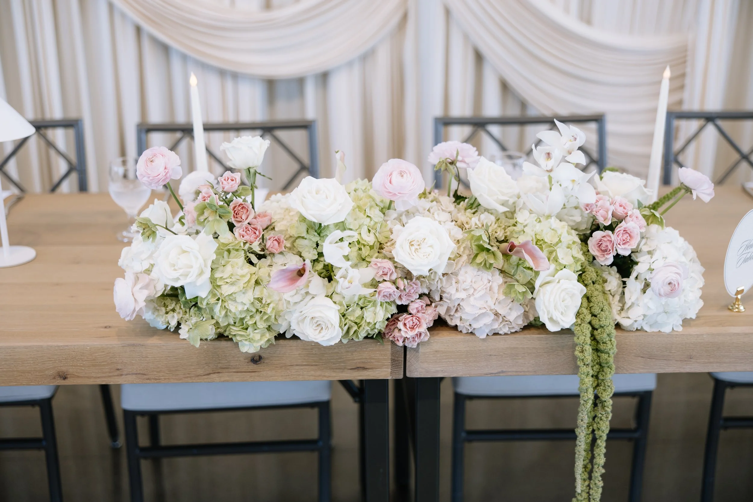 A lush floral arrangement of white, blush pink, and soft green blooms stretches across a wooden head table, accented by cascading greenery and tall white taper candles. Soft ivory draping creates an elegant backdrop behind the modern chairs.