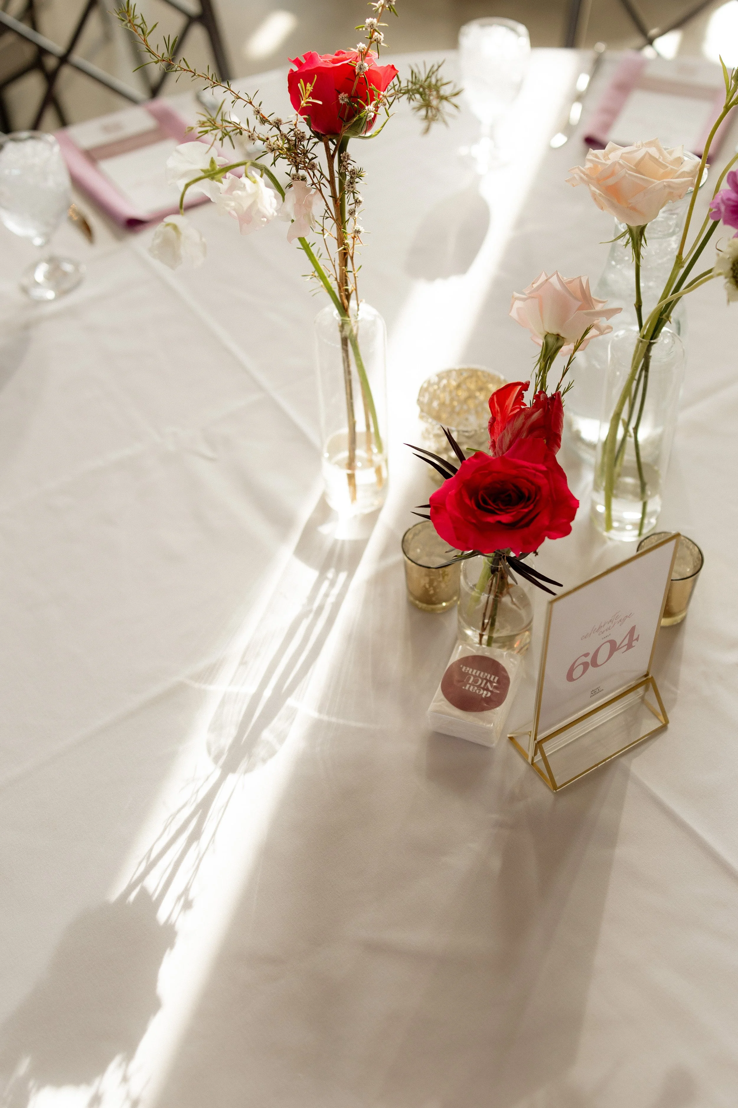 A table decorated with vases of pink and red roses and other flowers, with a gold table number stand displaying the number 604, set for a wedding or celebration.