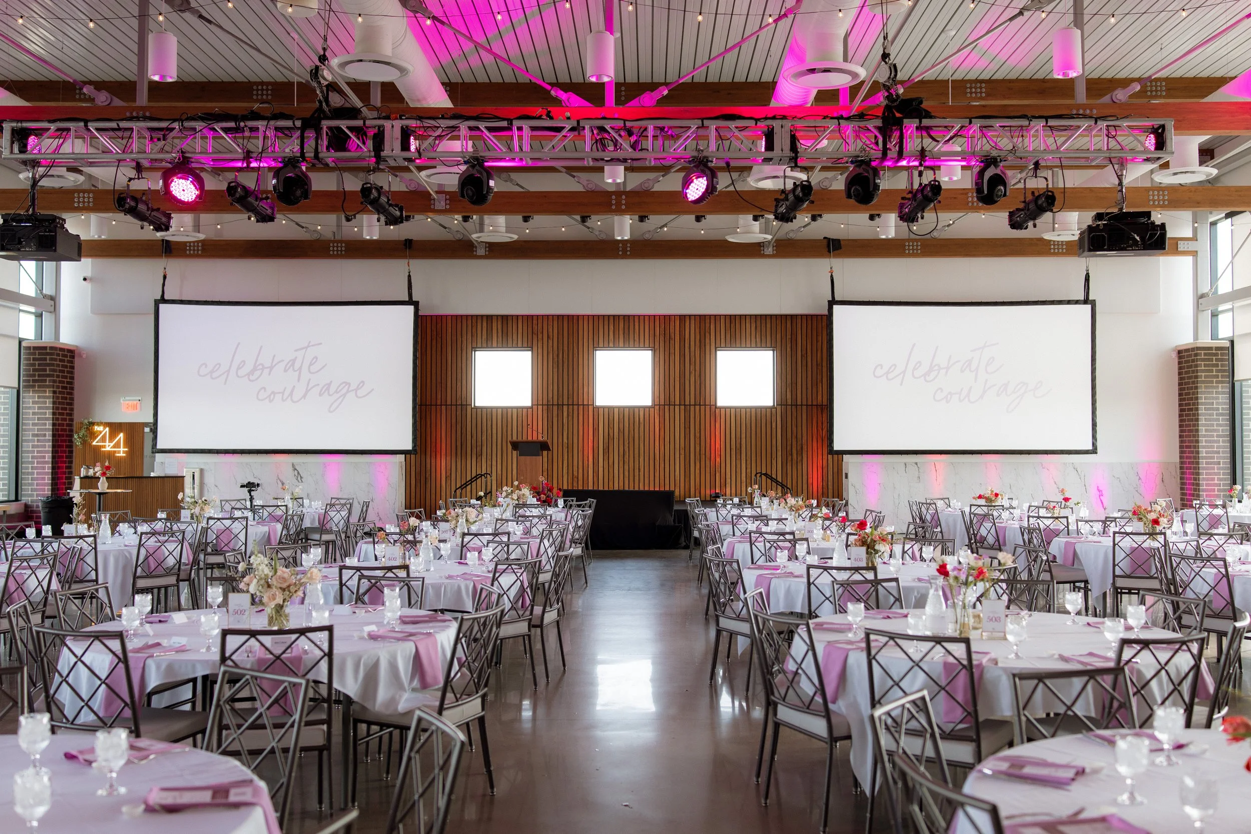 Large event hall decorated for a celebration with round tables covered in white tablecloths, pink napkins, and floral centerpieces. Two large screens display the words 'celebrate courage' on a wooden wall, and pink lighting illuminates the ceiling.