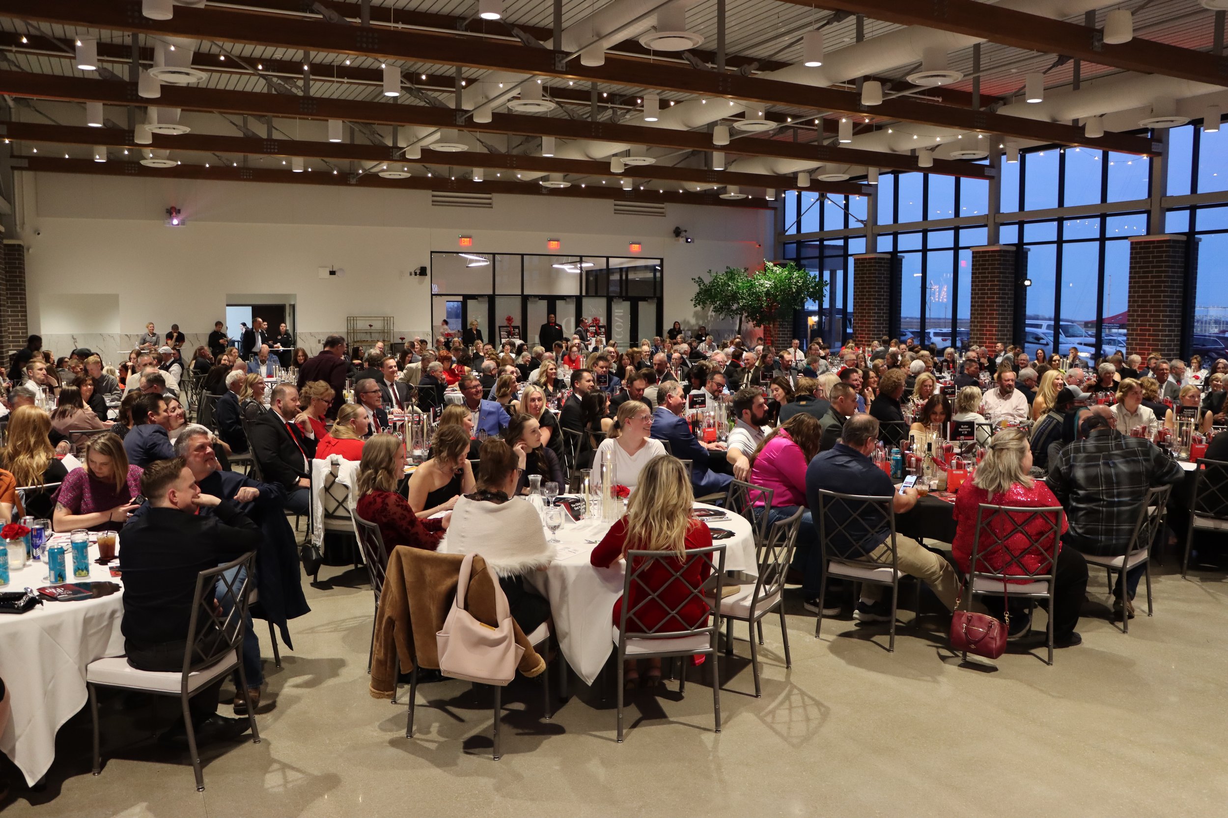 Large indoor gathering with many seated people at round tables during an event or celebration.
