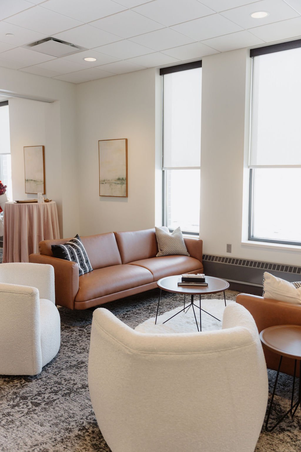 Modern living room with a tan leather sofa, white armchairs, a round black coffee table, and abstract artwork on the wall. Large windows with roller shades let in natural light.