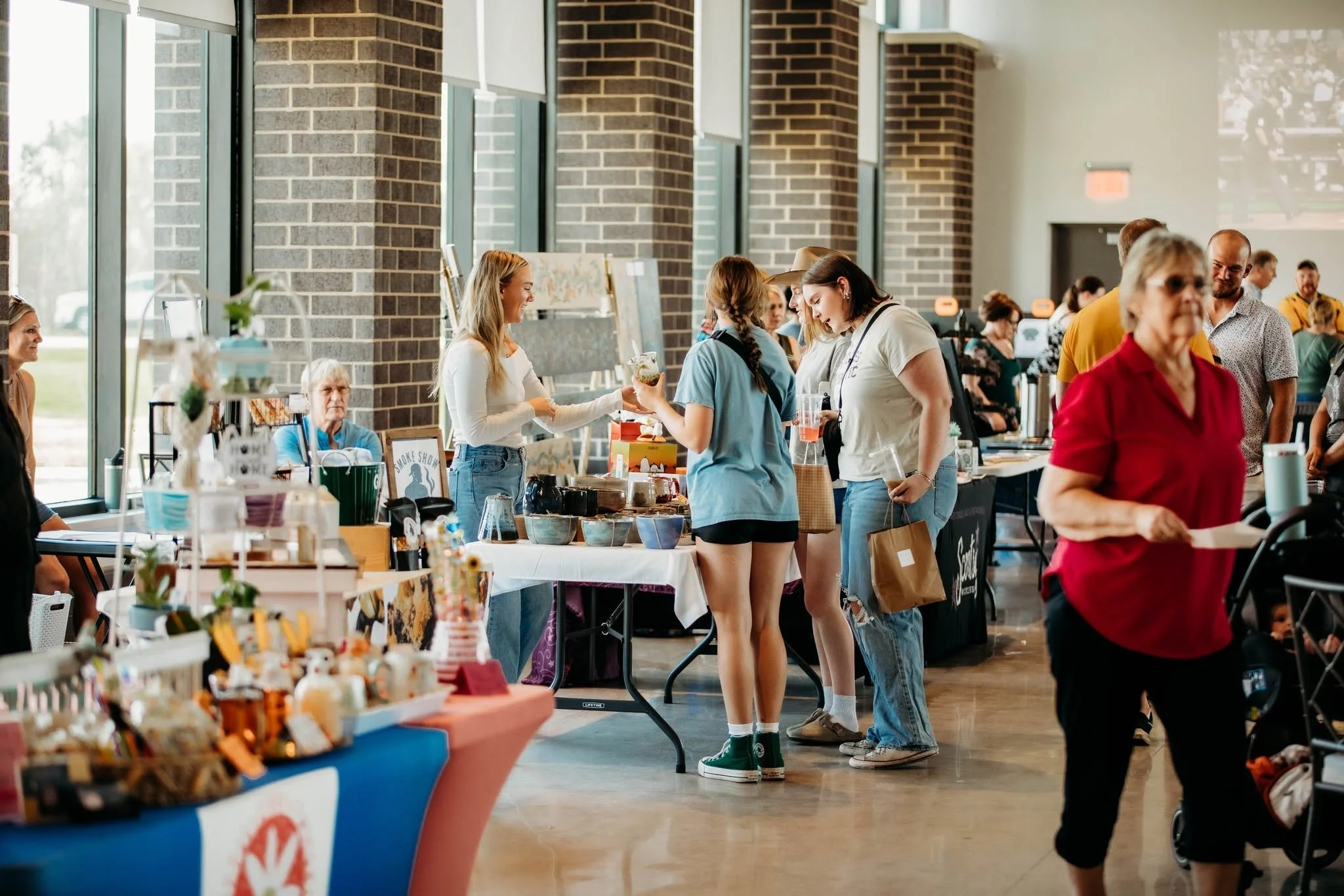 People shopping at an indoor market with tables of goods, brick wall with large windows, and a high ceiling.