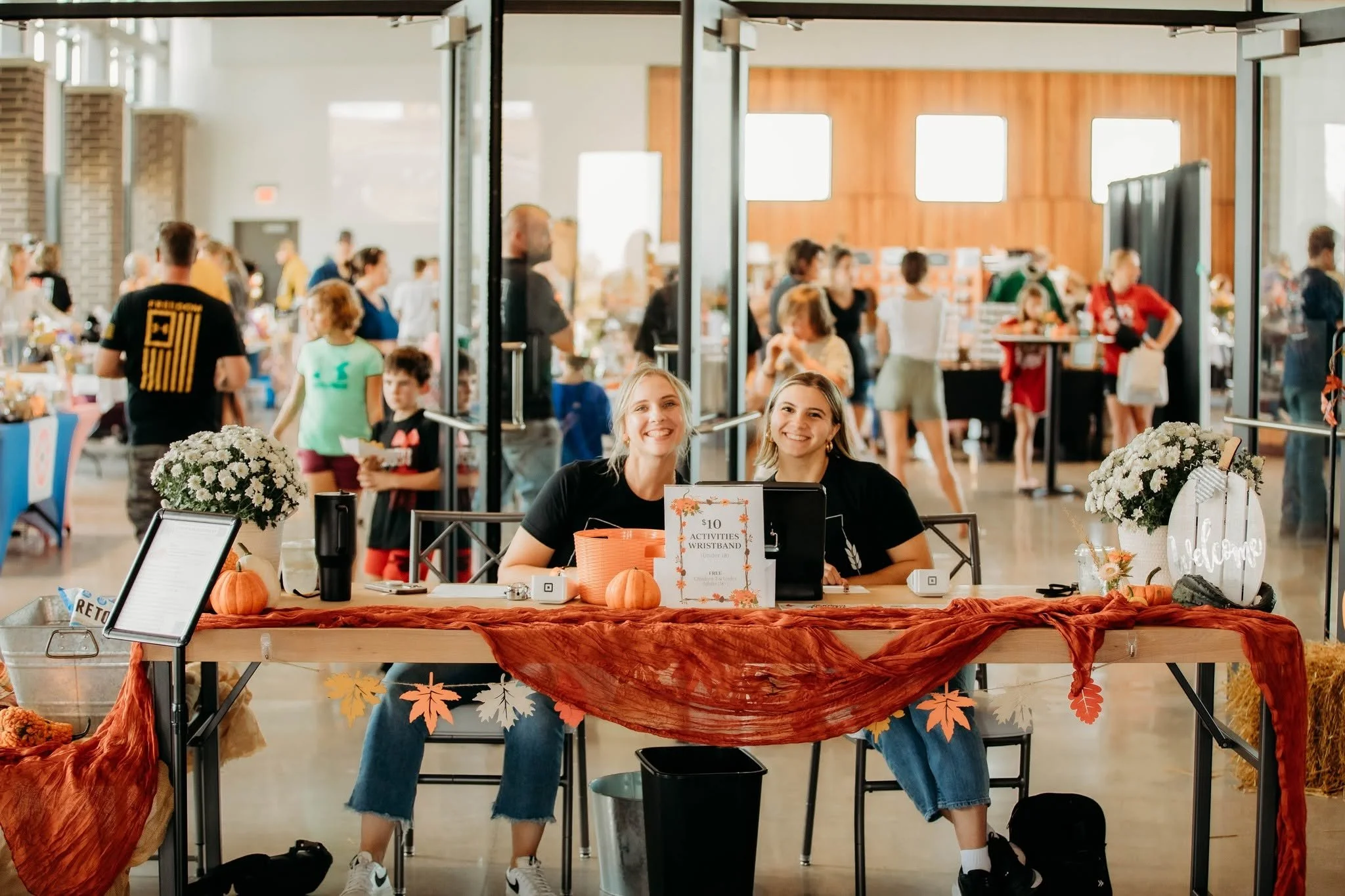 Two smiling women sitting at a registration table decorated with autumn-themed items, including pumpkins and fall leaves, inside a busy event space with people walking and browsing in the background.