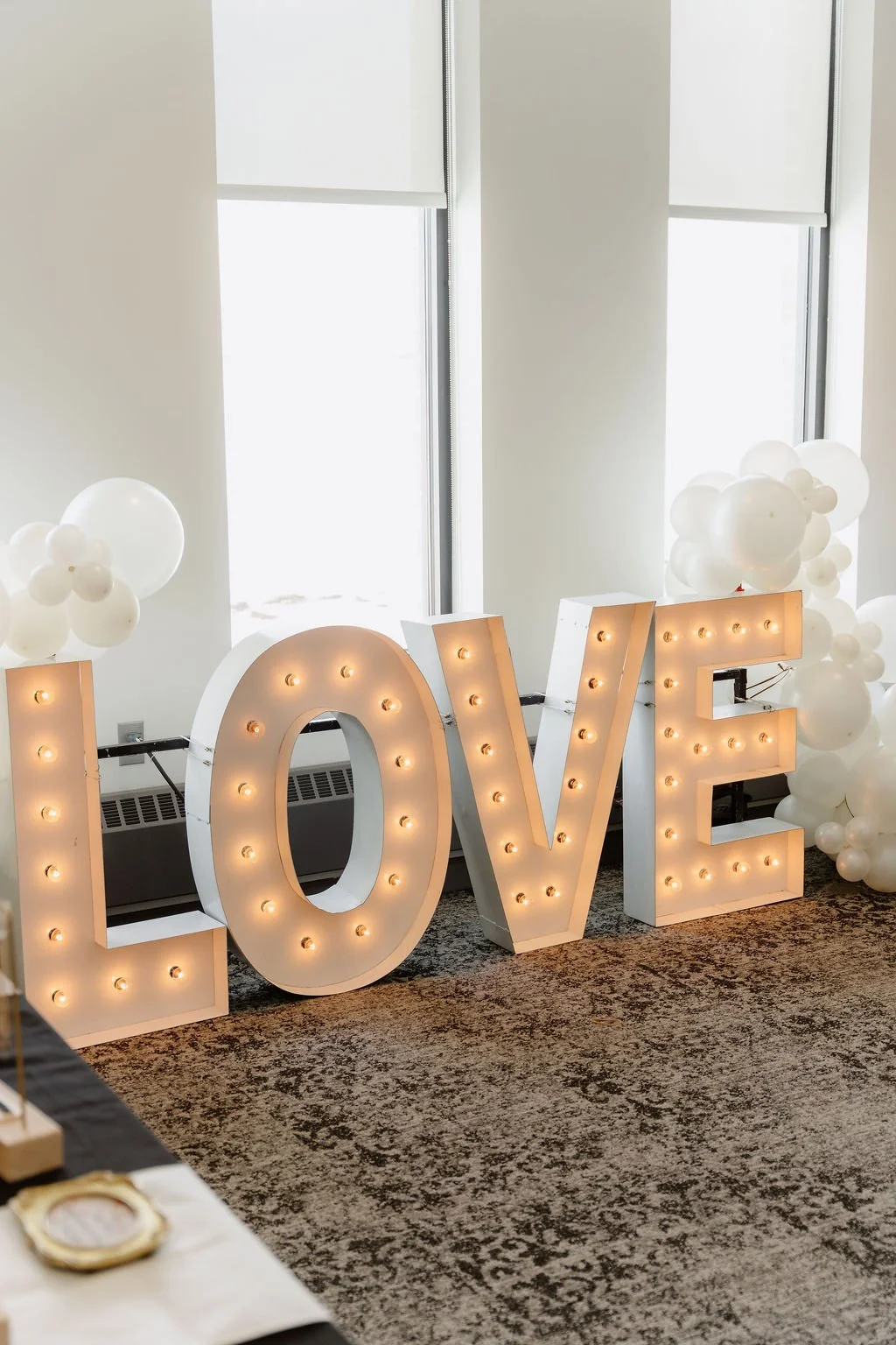 Large illuminated letters spelling "LOVE" with white balloons in the background, inside a bright room with tall windows.