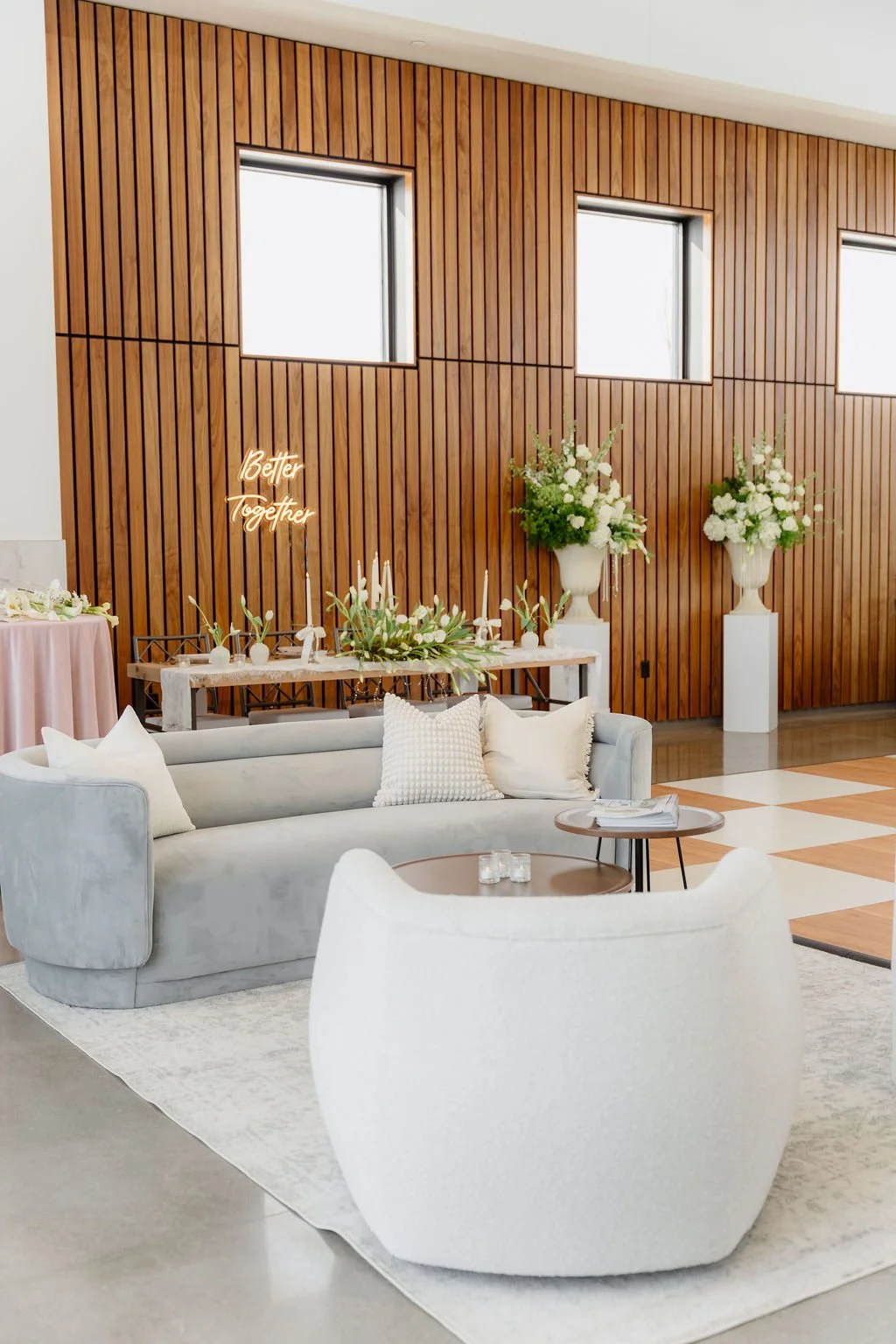 Modern event setup with a light gray sofa, white armchair, and small round tables in front of a wooden-paneled wall. The wall features a neon sign saying "Better Together" and two tall white vases with white flowers. A decorated table with floral arr