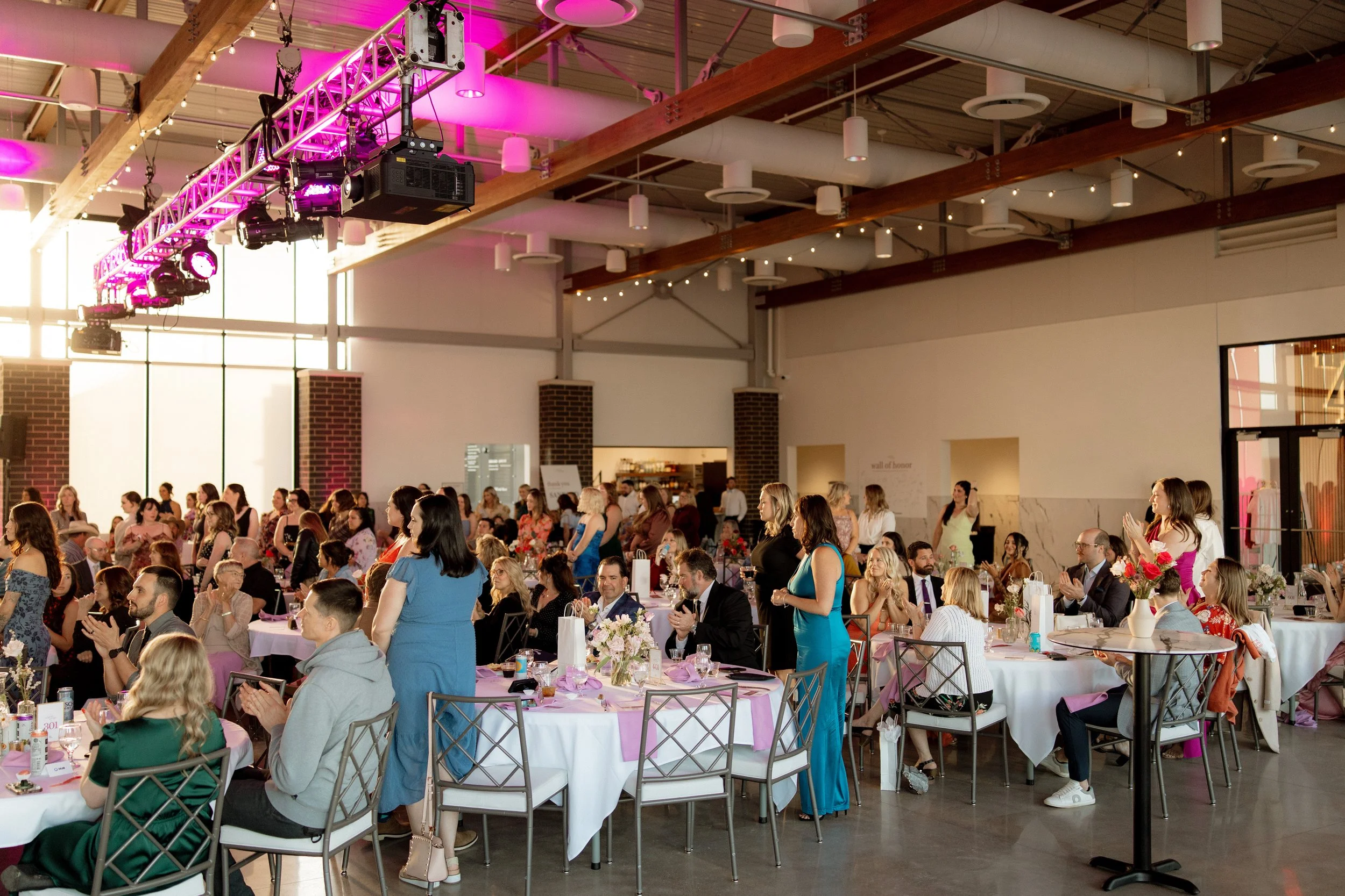 A large indoor event with many people seated at decorated tables, some standing and clapping. There are pink and white decorations, flowers on tables, and pink lighting accents on the ceiling. The space has high ceilings with exposed wooden beams and