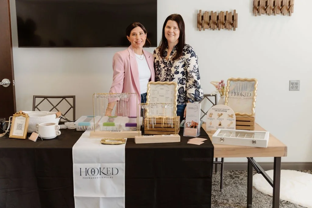 Two women standing behind a table display at a jewelry event, with jewelry items, signs, and promotional materials on the table, and decorative wall art in the background.