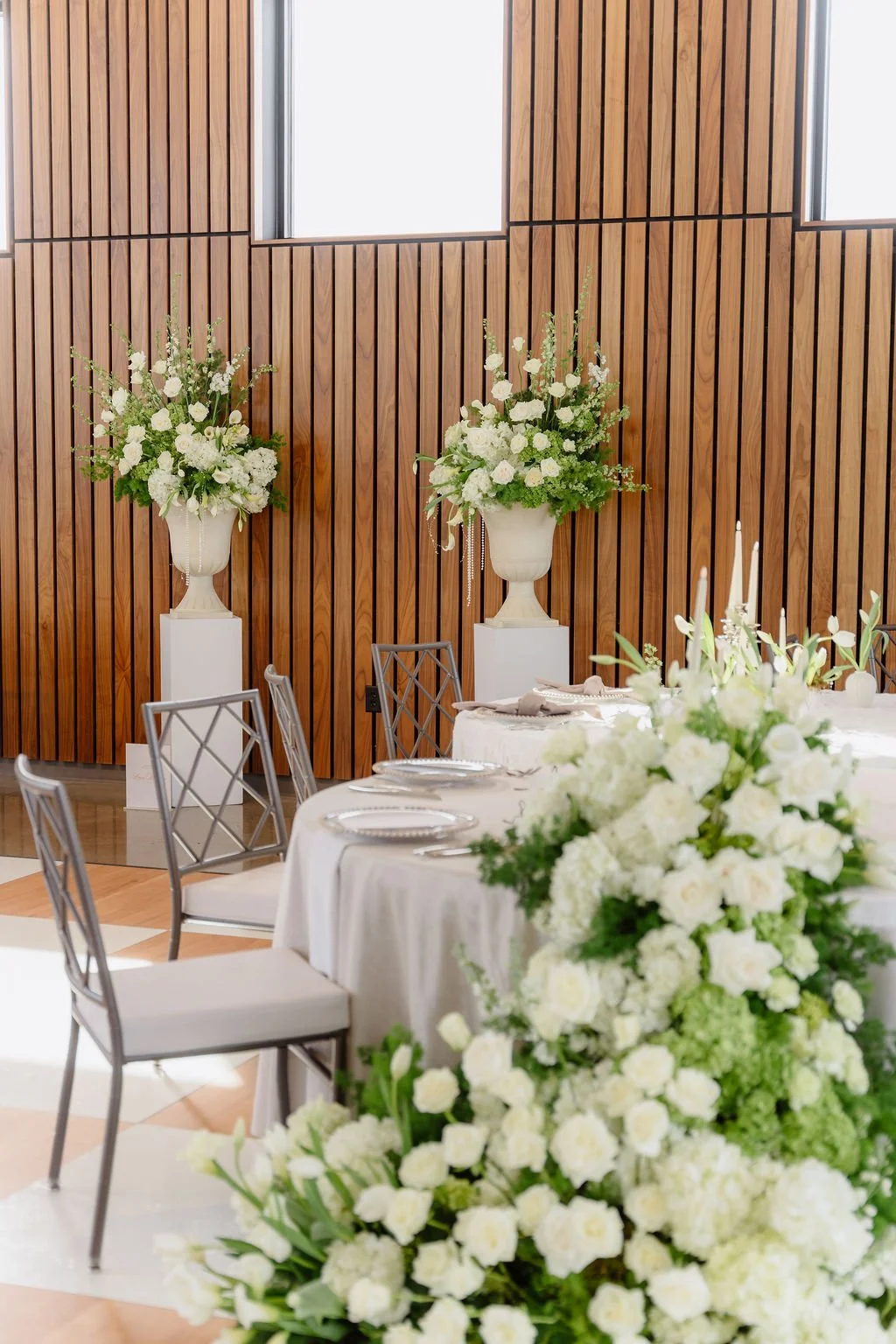Elegant event space with white floral arrangements on white pedestals and a long table set with white flowers and candles, against a wood-paneled wall with high windows.